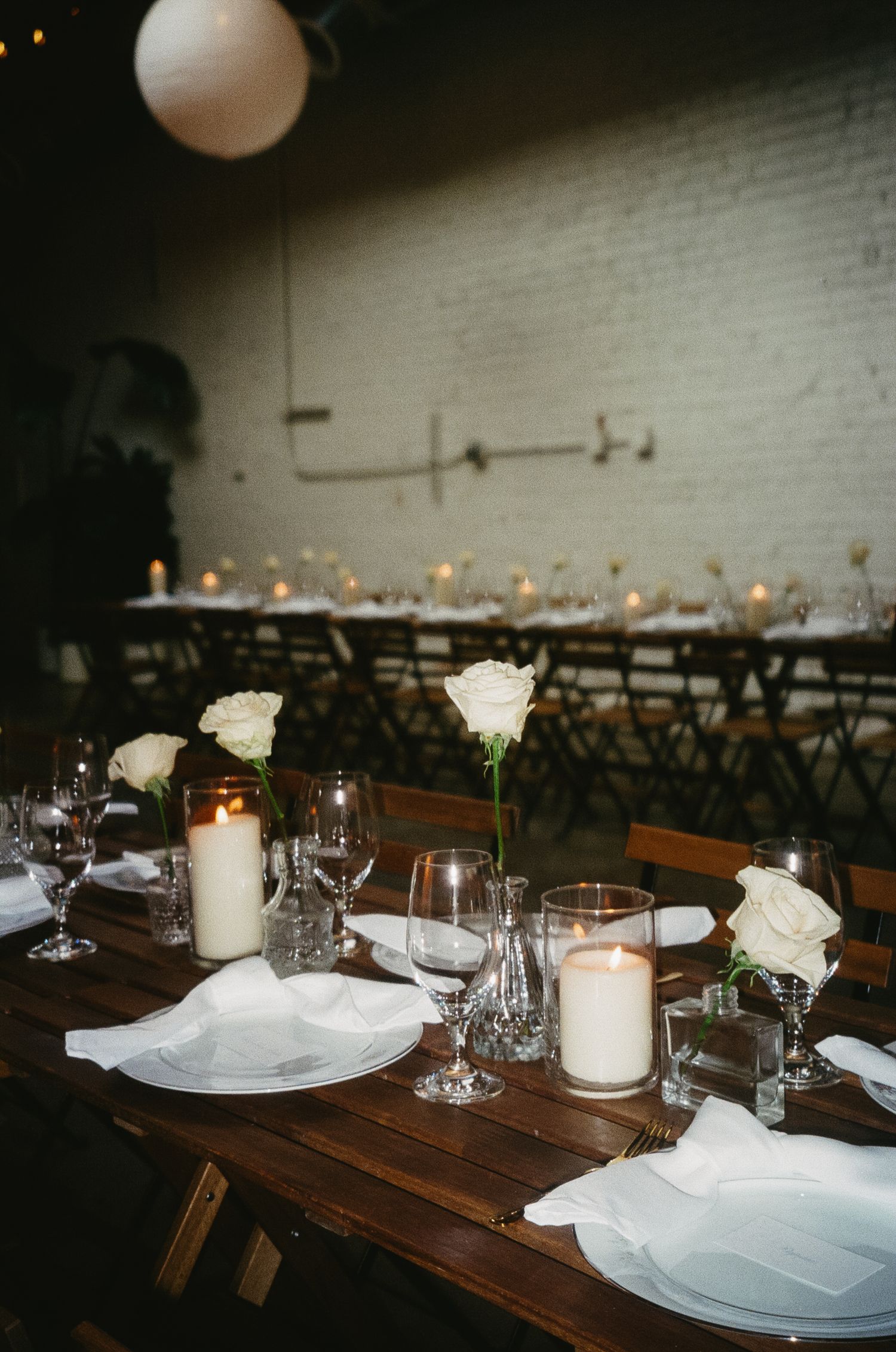 Elegant table setting with white roses, candles and wine glasses on rustic wooden tables in a moody indoor venue.