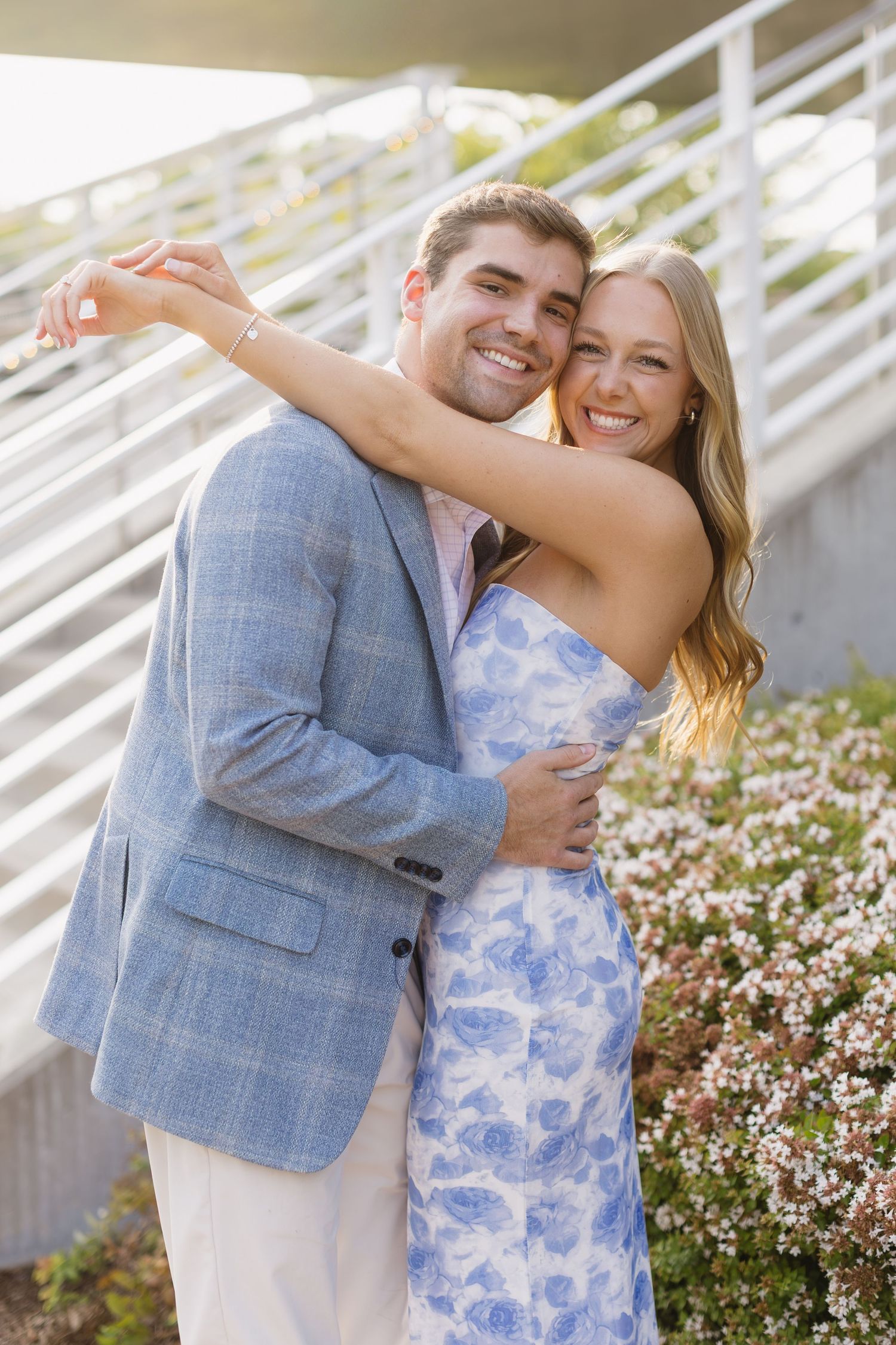 A couple embraces while standing near flowering bushes, wearing coordinated blue and white summer outfits.