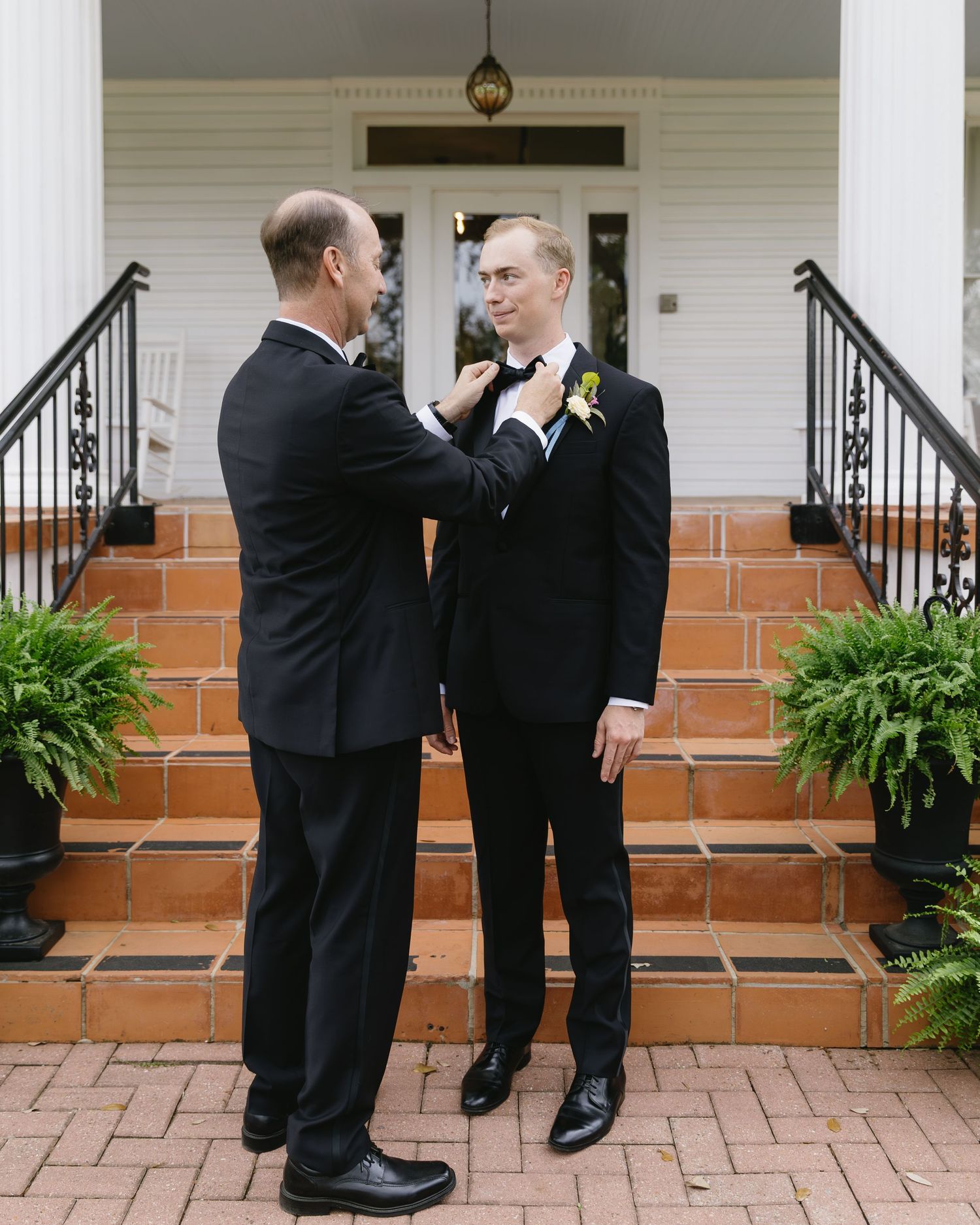 Two people in formal attire sharing an intimate moment on brick steps with decorative ferns on the porch.