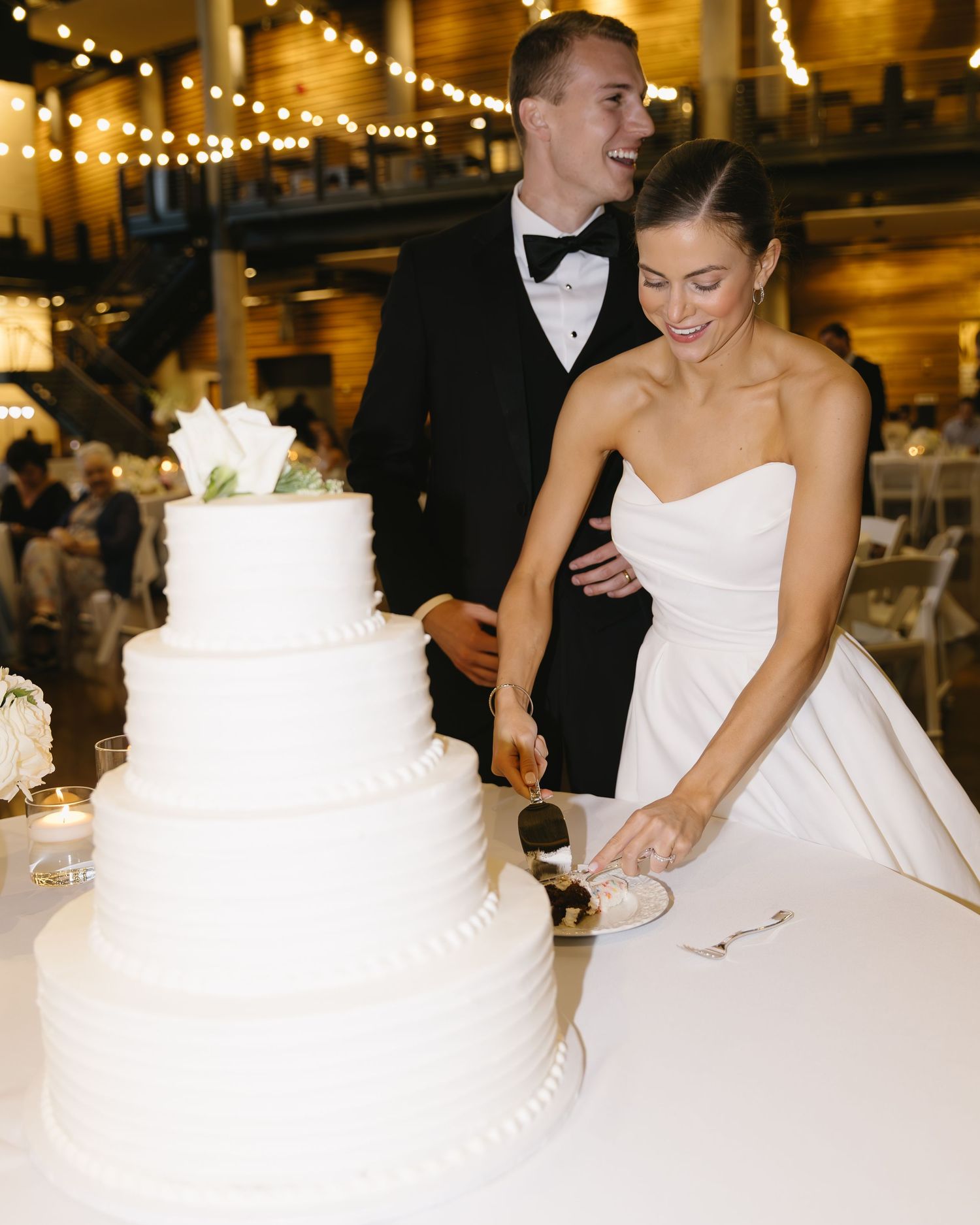 Wedding couple cutting their white tiered cake at reception under string lights.