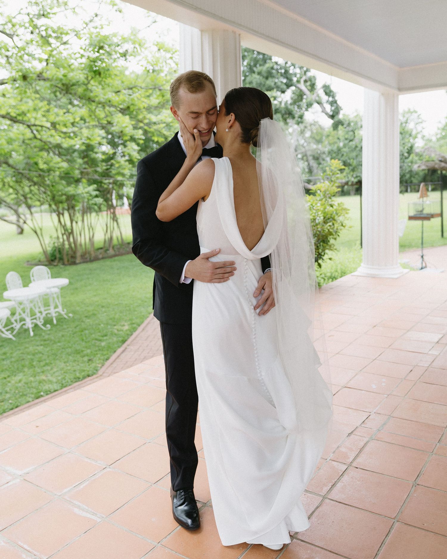 A romantic wedding couple shares an intimate moment on a covered porch with lush greenery in the background.