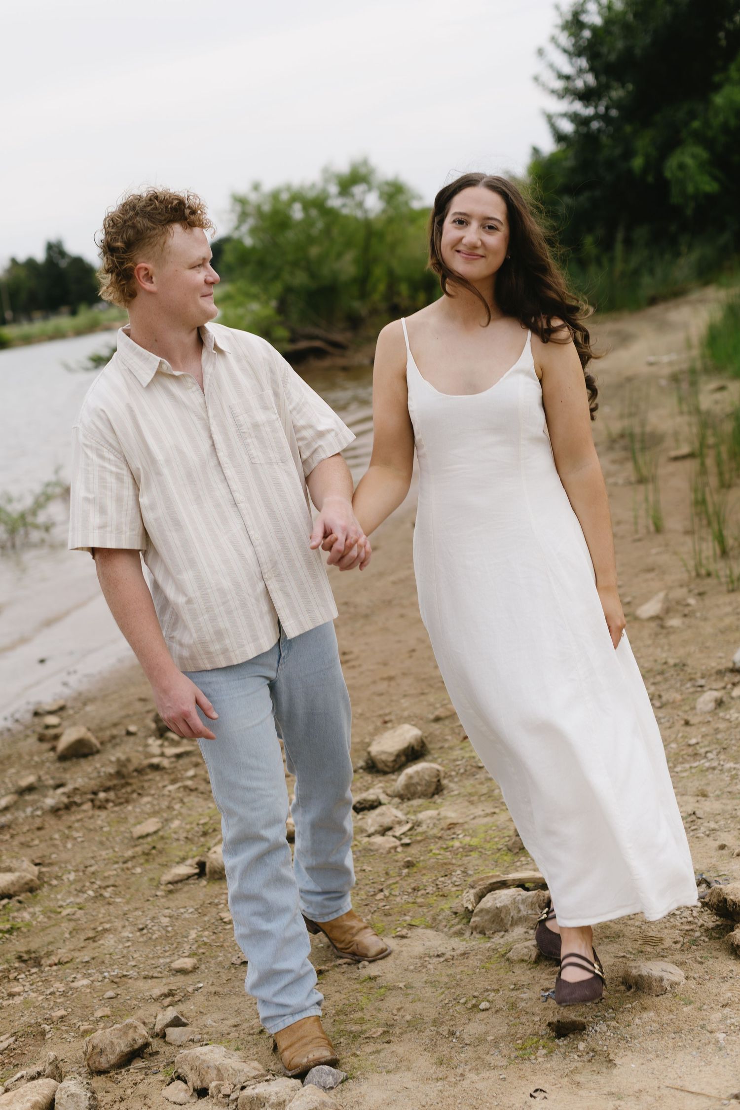 A couple explores a rocky beach path together wearing matching white and denim outfits.