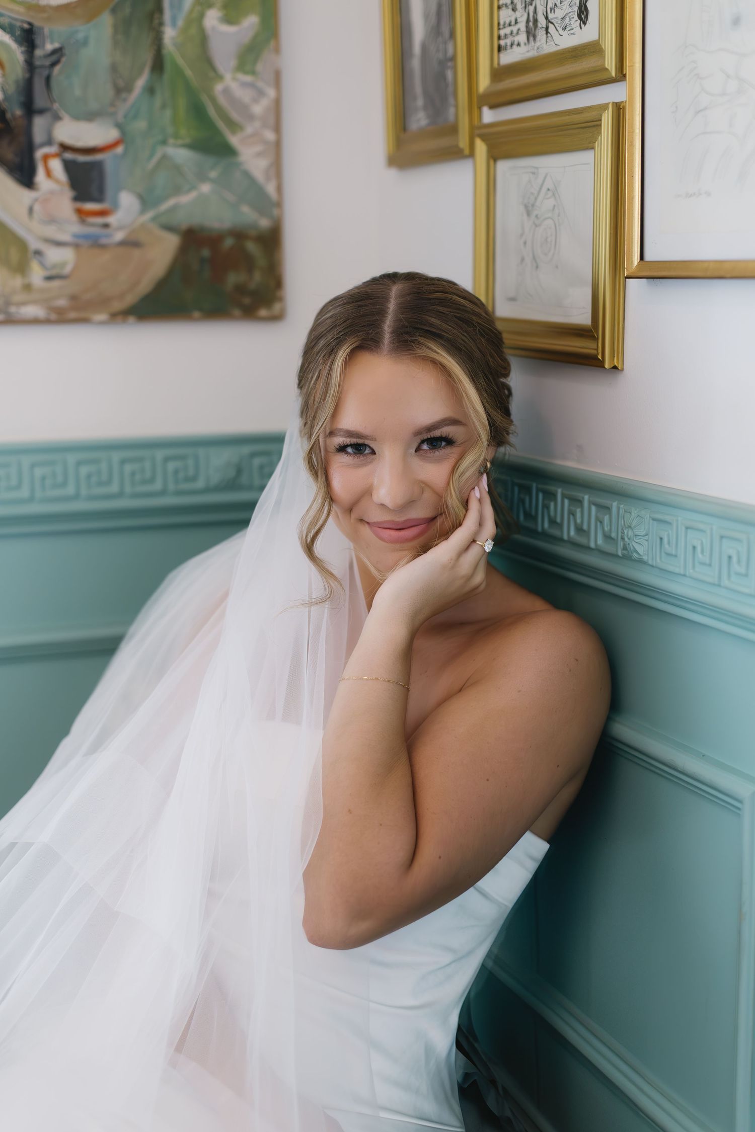 A bride in an elegant white gown rests her head on her hands while seated against turquoise wainscoting.