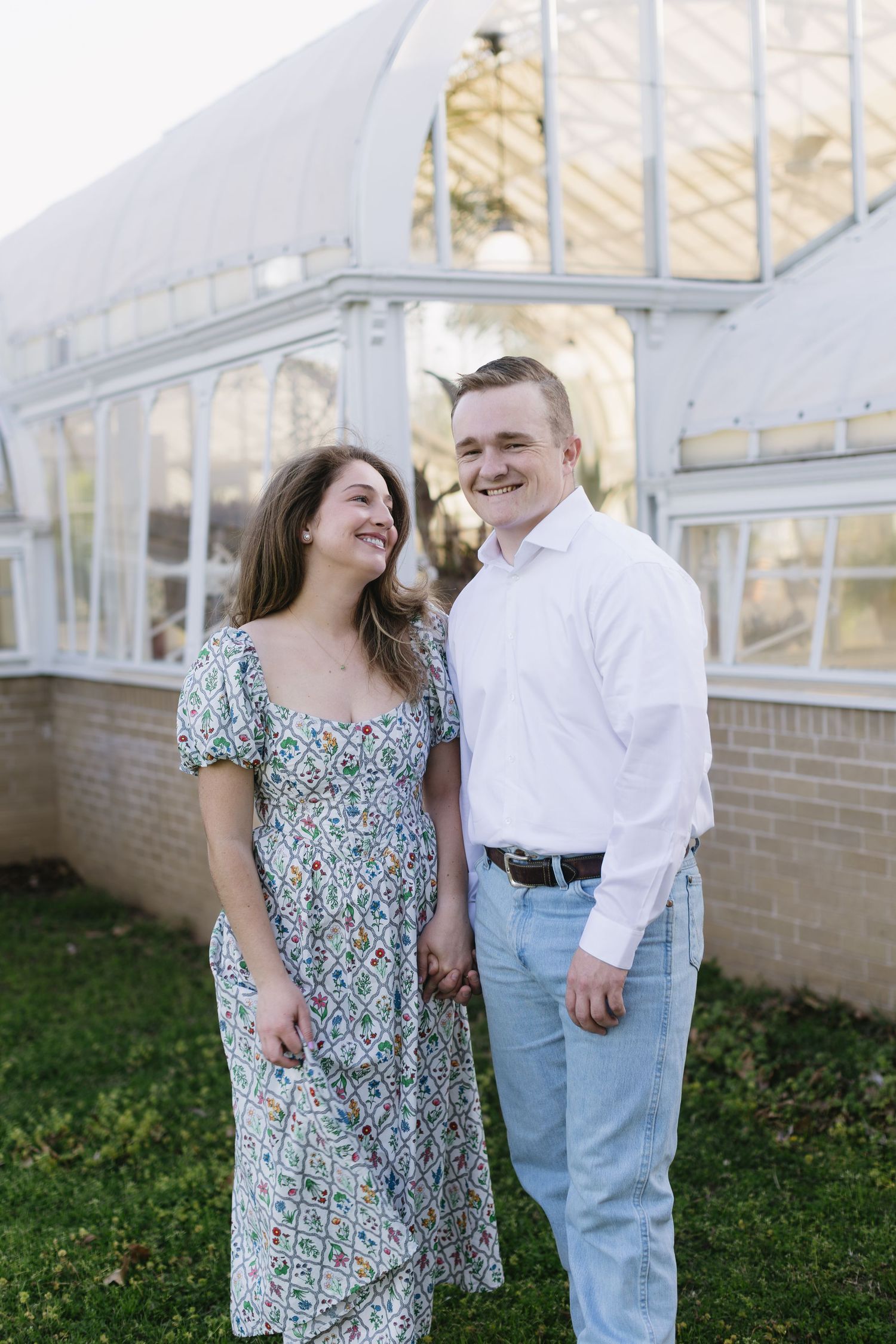 A couple stands together near a greenhouse, sharing a tender moment in natural lighting.