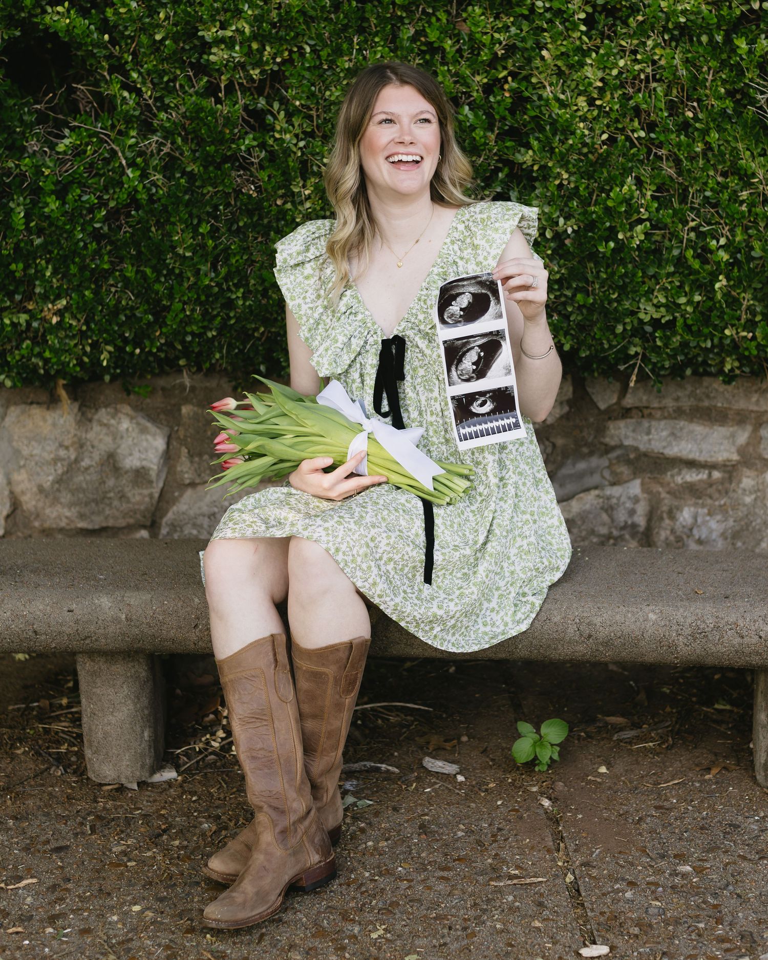 Person in a floral dress and brown boots sits on a stone bench showing off ultrasound photos with joy and excitement.