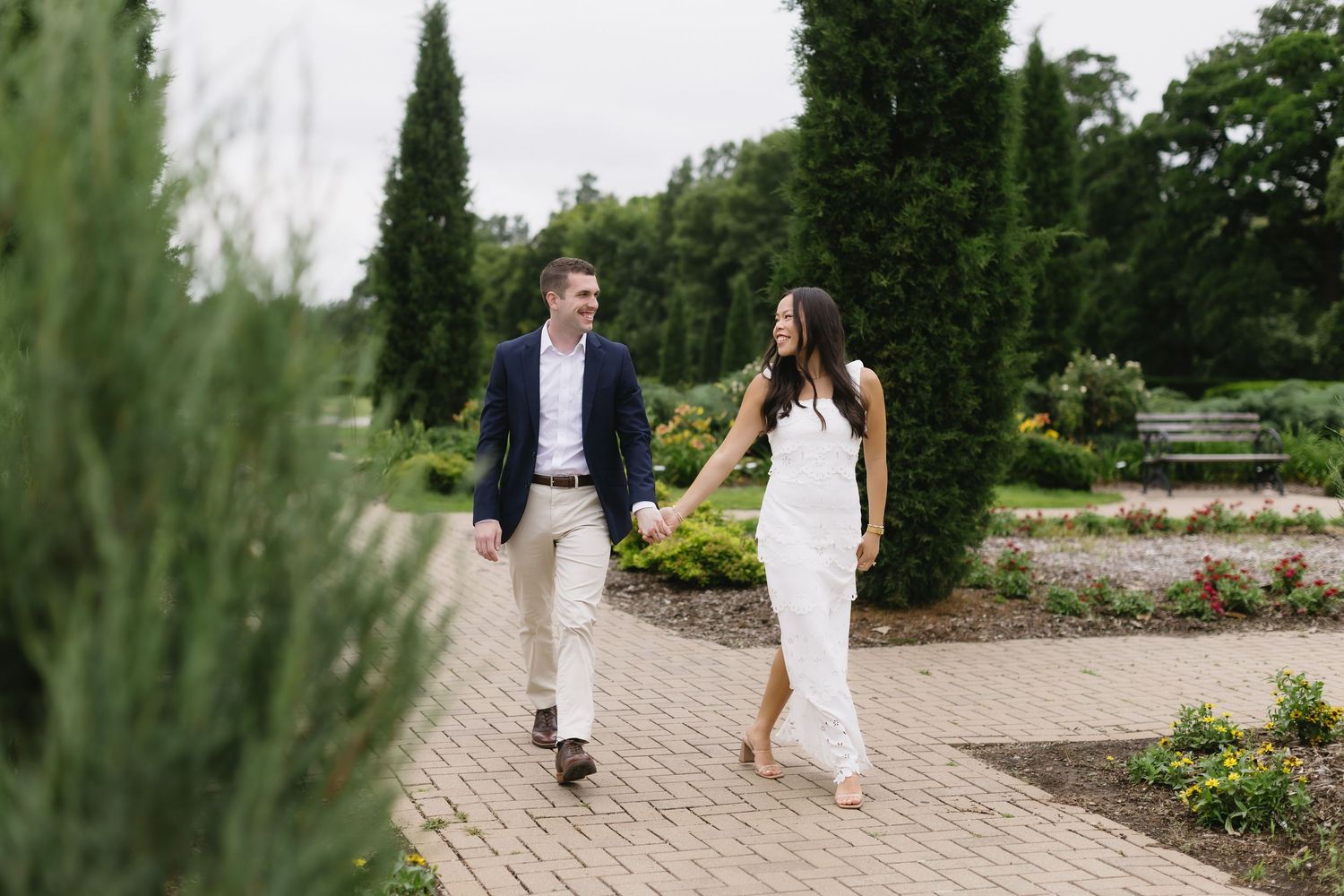 A couple in light summer clothing walks along a garden path surrounded by lush landscaping and greenery.