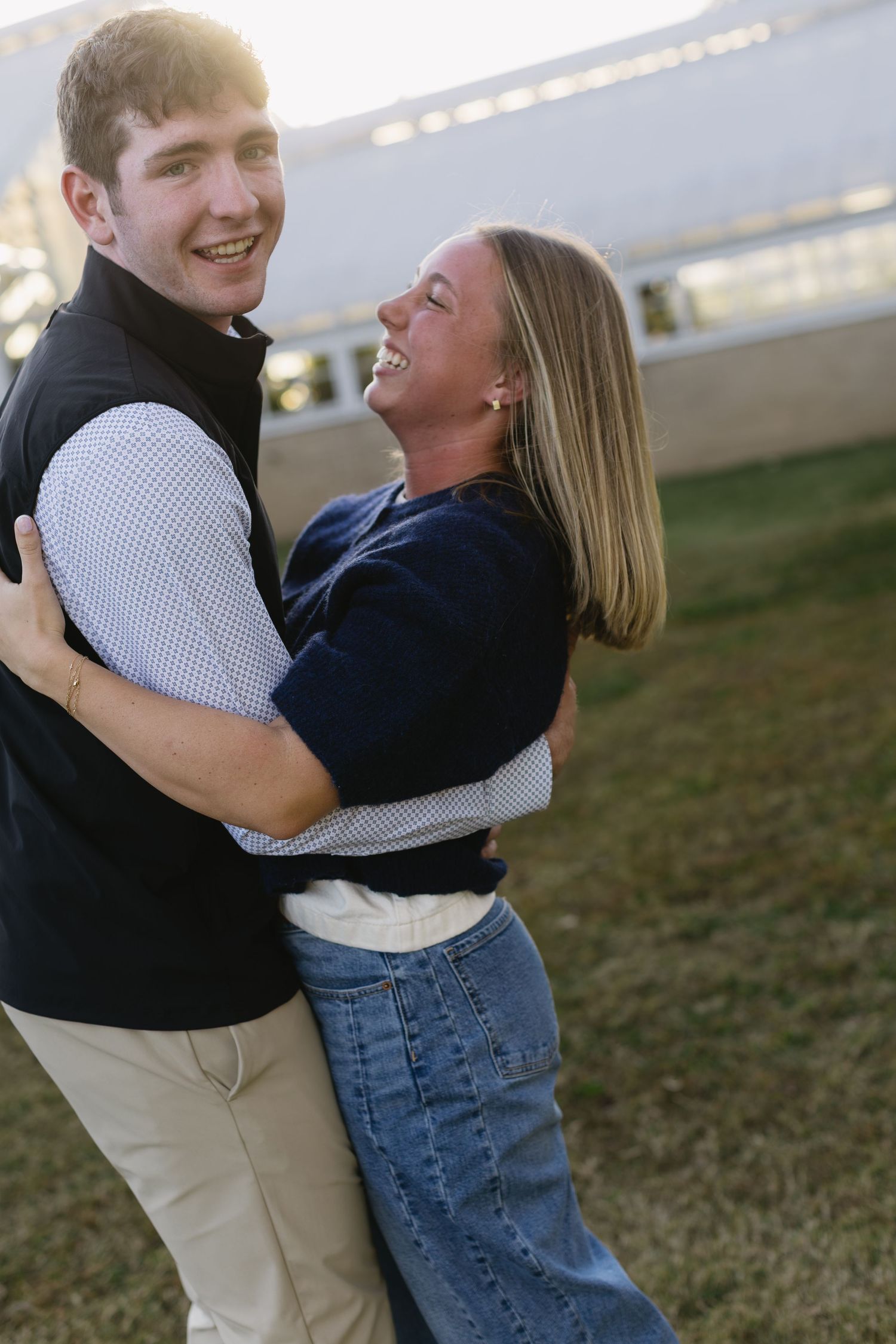 A couple laughs and embraces outdoors during a casual photoshoot at sunset.