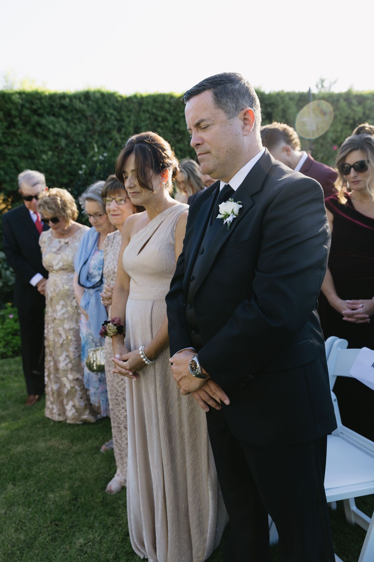 Wedding guests in formal attire gather on lawn during outdoor ceremony.
