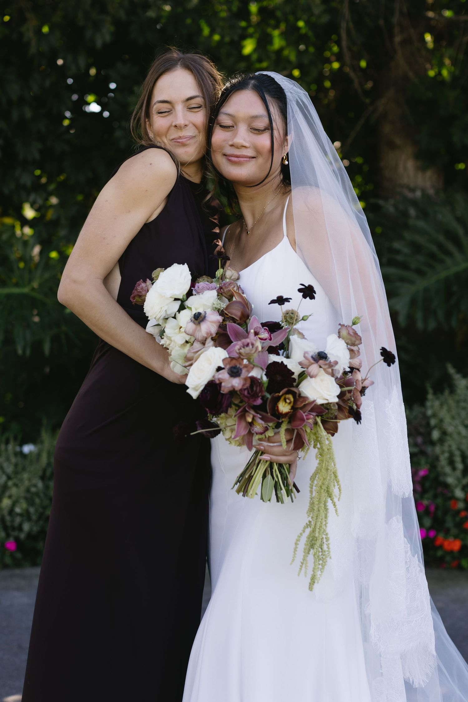 Bride in white dress embraces bridesmaid in black while holding purple and white floral bouquets outdoors.