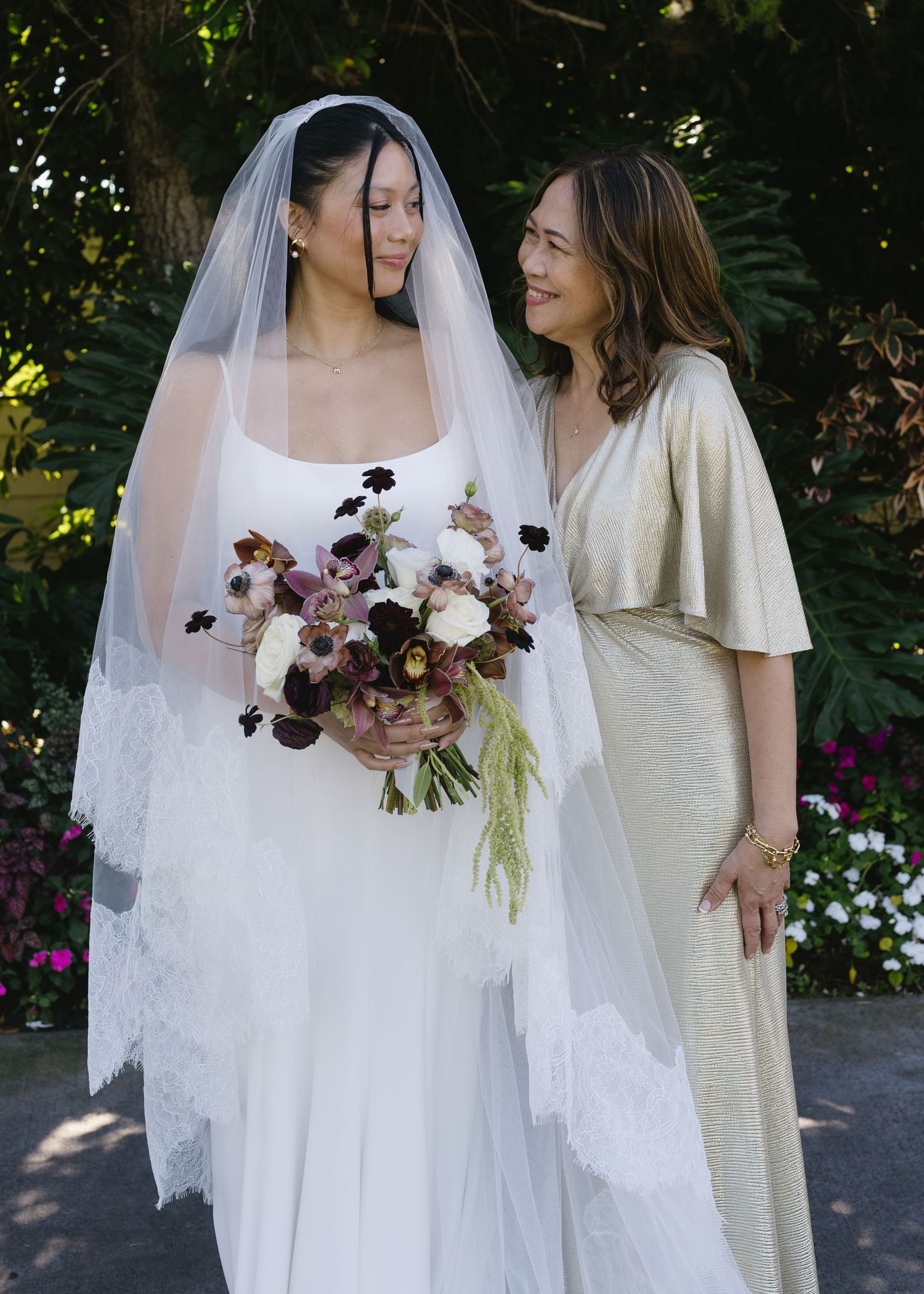 Bride in white gown holding bouquet while standing with companion in garden setting.