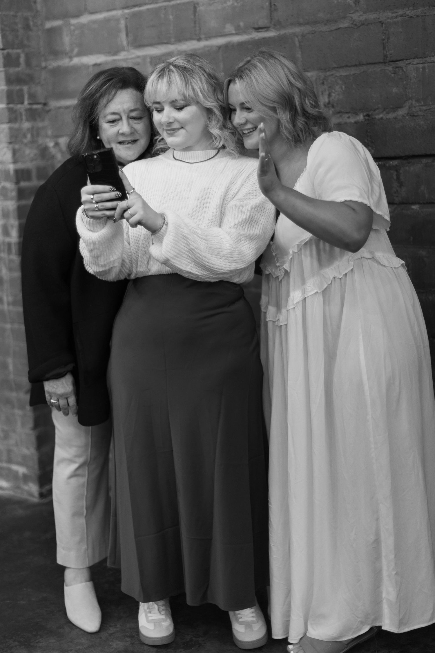 Three friends sharing a laugh while looking at a phone together against a brick wall in black and white.