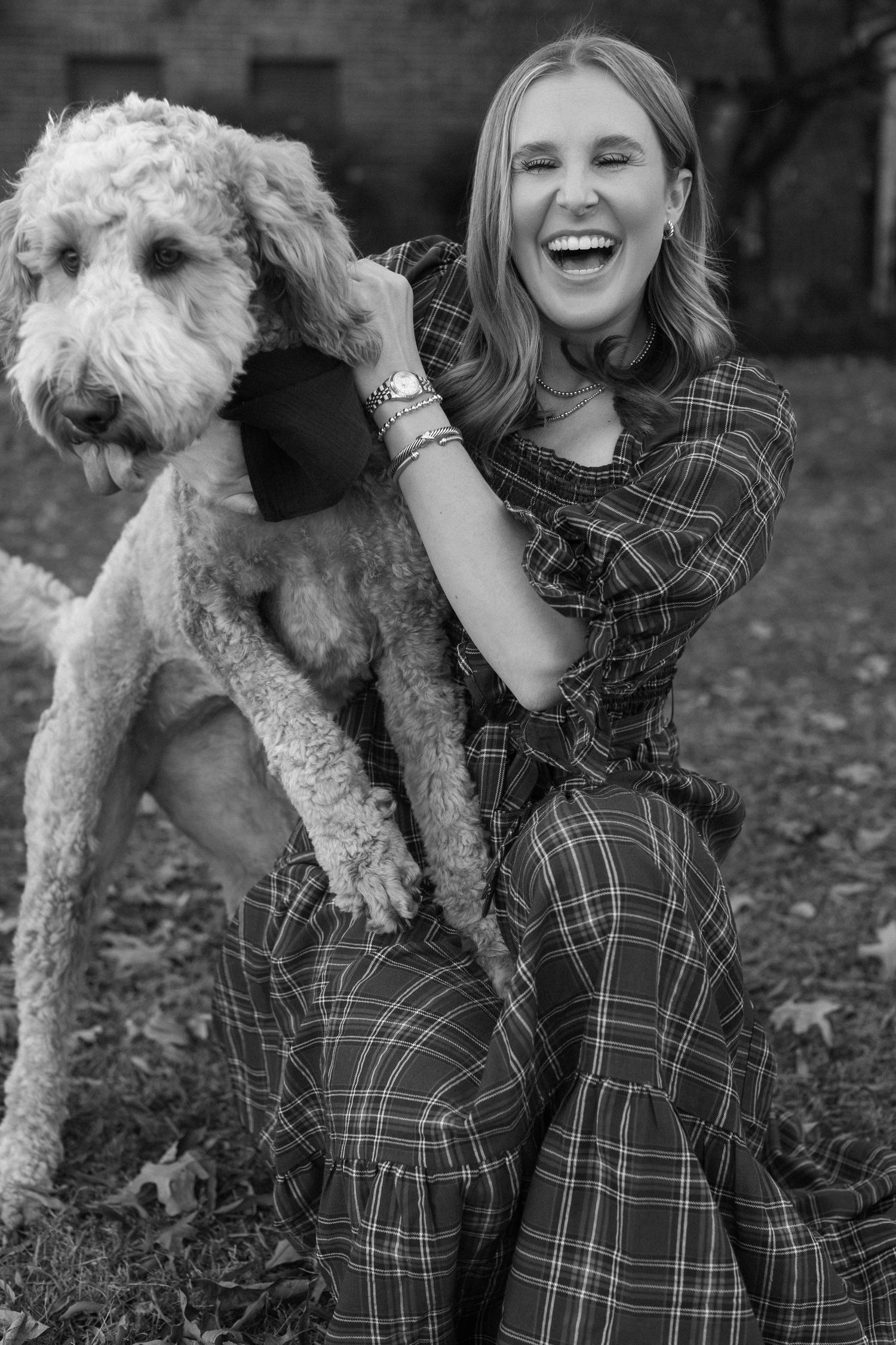 A goldendoodle poses playfully with its owner in a plaid dress during a black and white photoshoot.