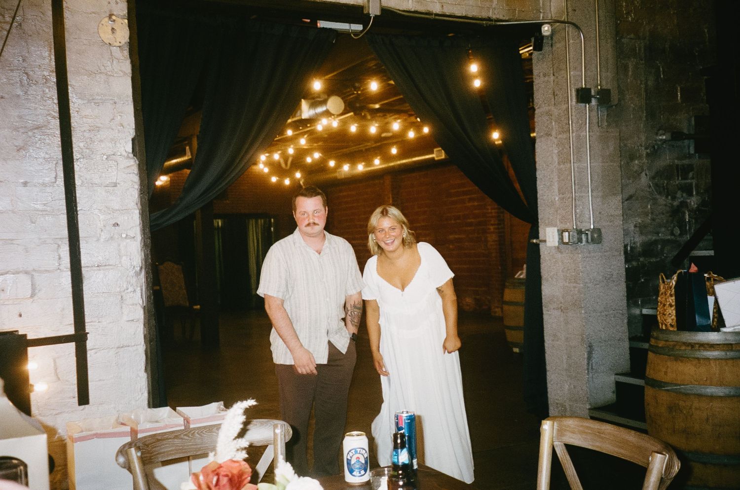 Indoor venue space with exposed brick walls, string lights, and dark curtains creating a rustic atmosphere.