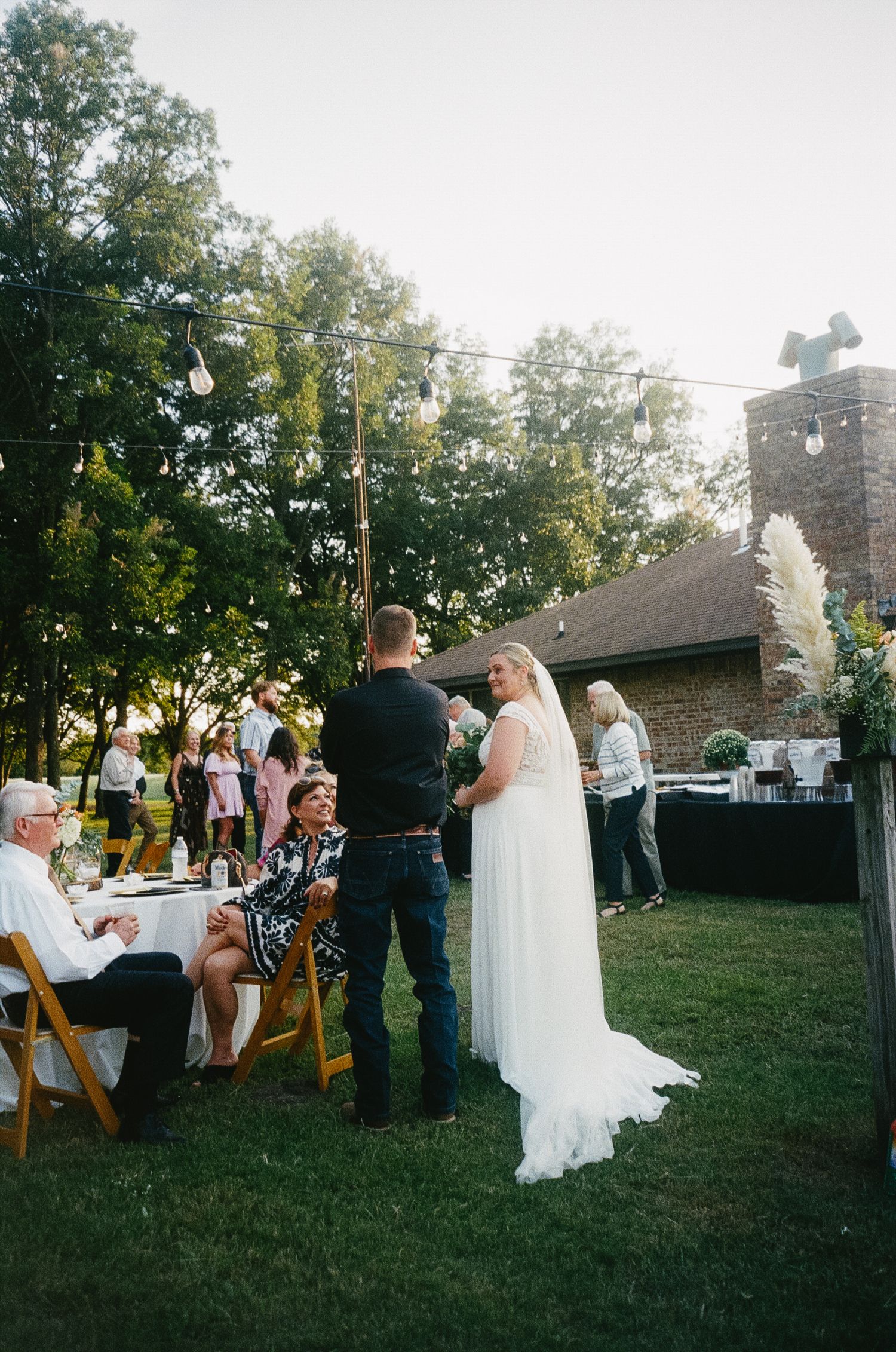 Outdoor wedding reception at sunset with guests seated at tables while couple stands on grass near stone building.