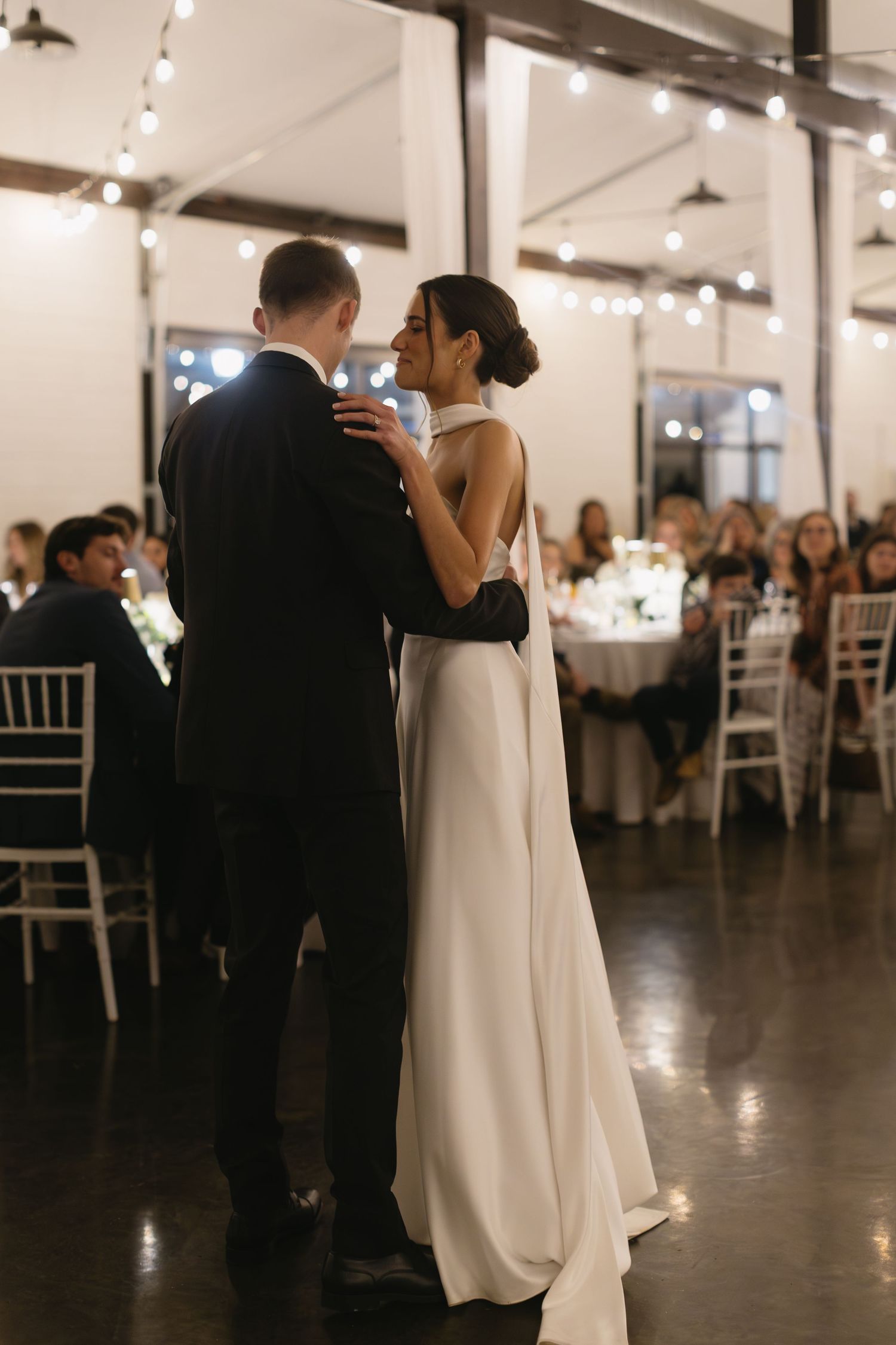 A couple shares their first dance in an industrial-style wedding venue with string lights overhead.