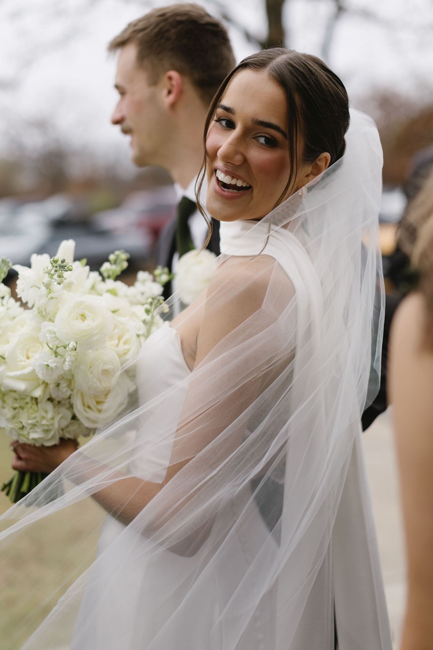 Elegant bride with flowing veil and white bouquet smiles during wedding celebrations.