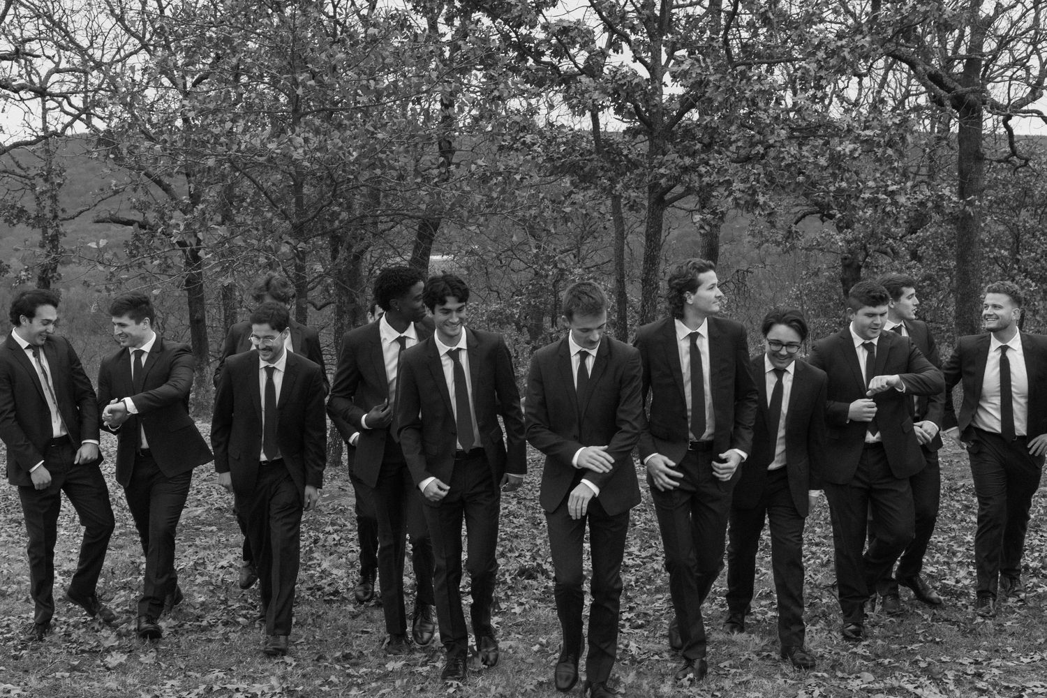 A group of groomsmen in black suits walk together through an autumn forest landscape.