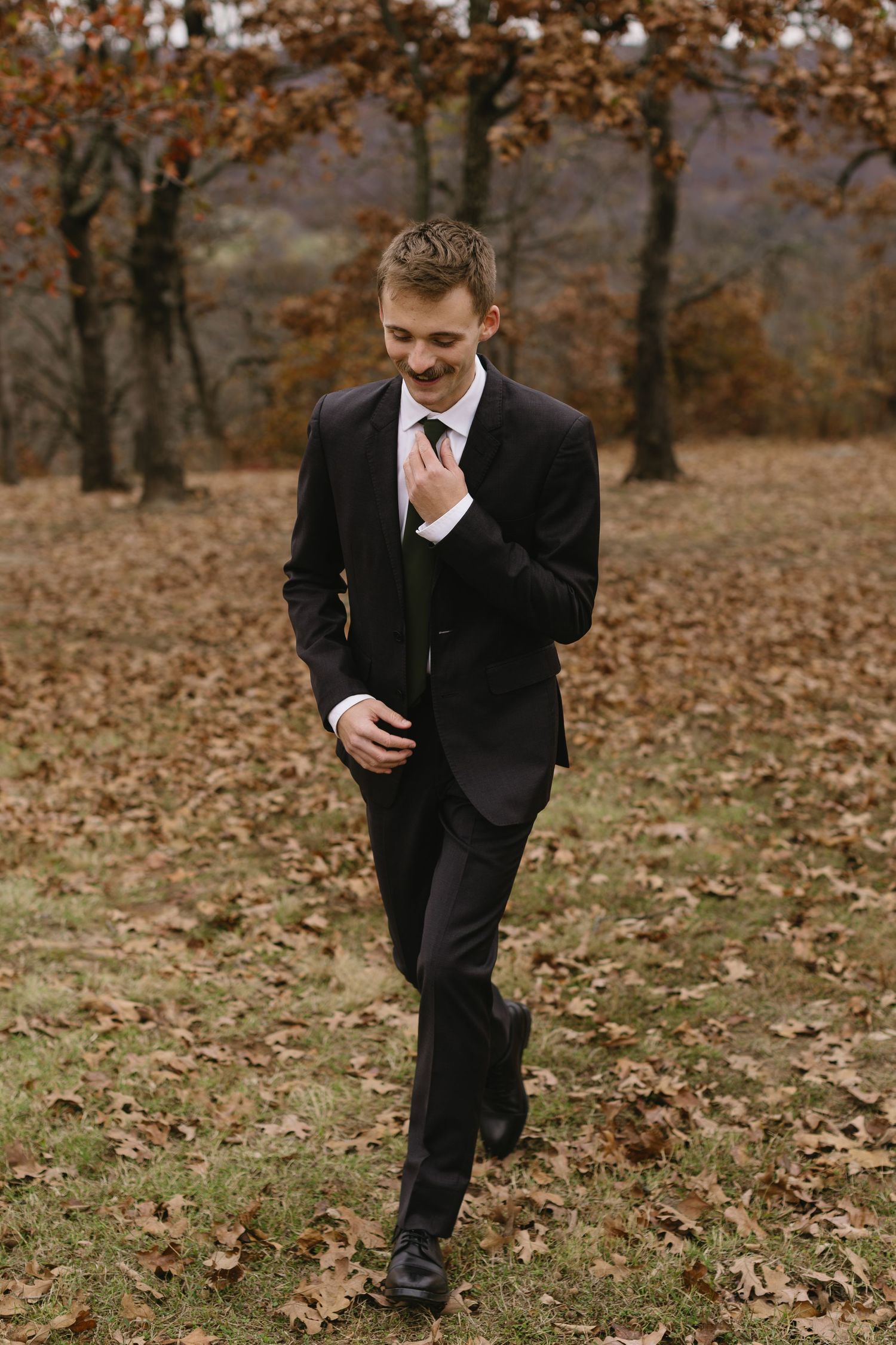 A groom in a classic black suit walks through fallen autumn leaves in a wooded setting.