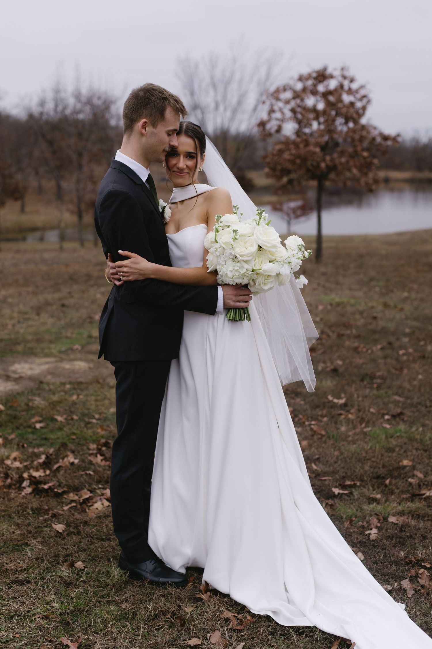 A romantic outdoor wedding portrait with a white bridal gown and bouquet against an autumn lakeside backdrop.