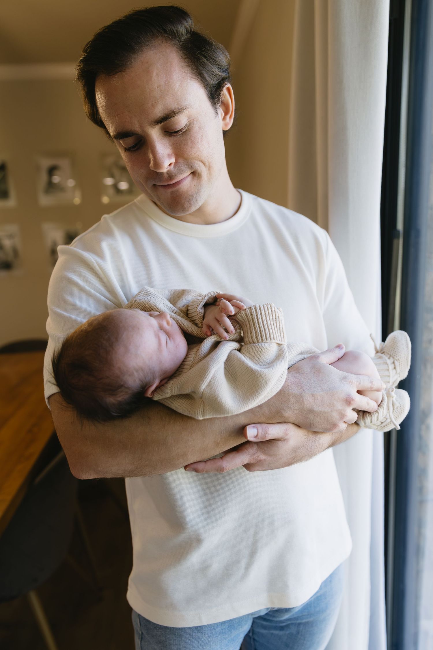 Parent in white shirt holding and bonding with infant near window.