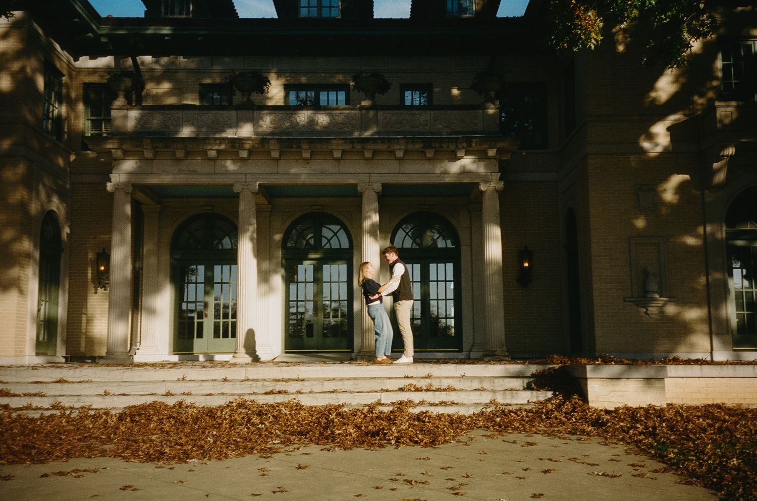 A couple sits on stone steps in front of an elegant historic building during an autumn day.