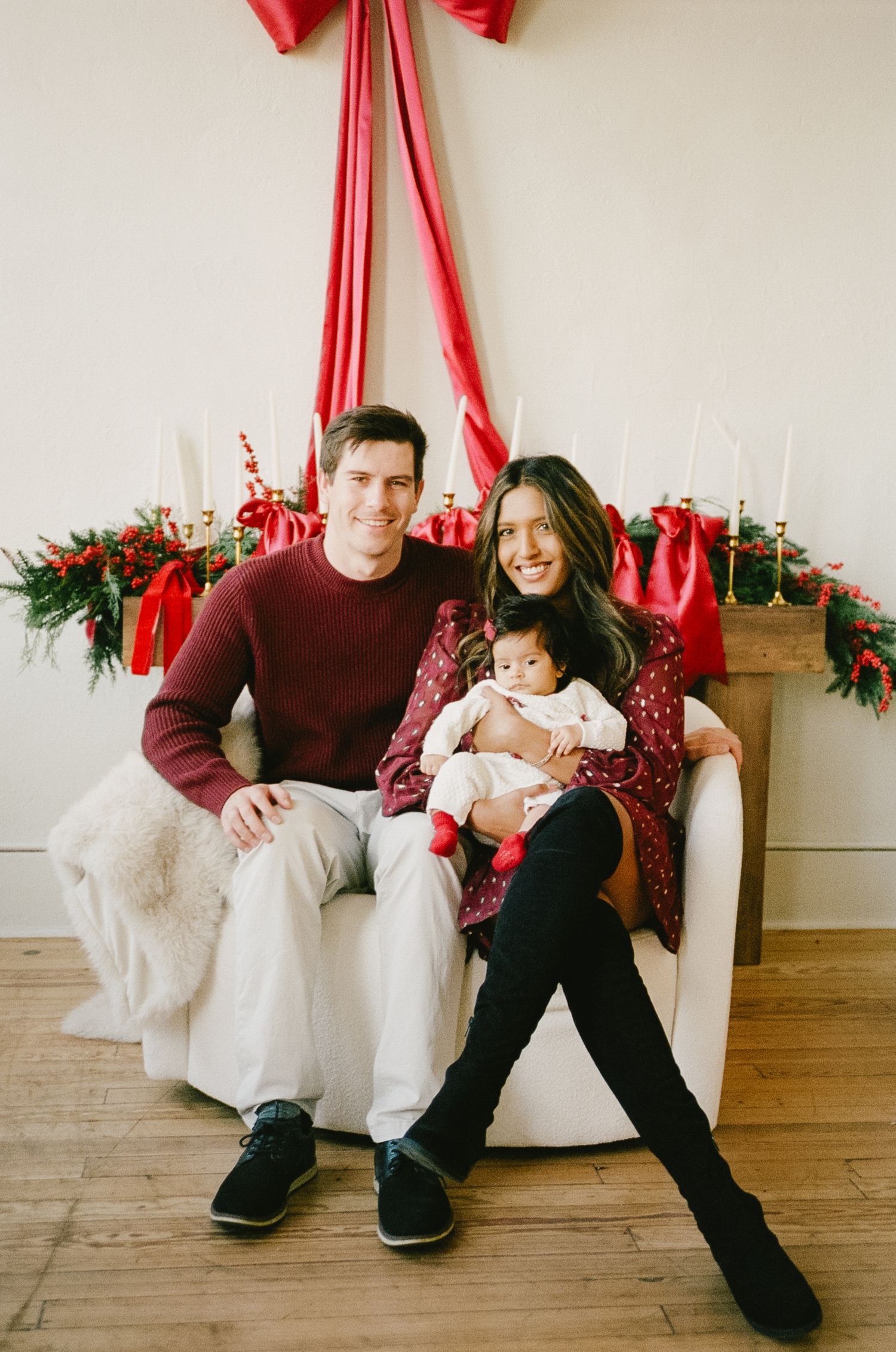 A couple sits together on a white chair with festive red ribbons and holiday decorations in the background.