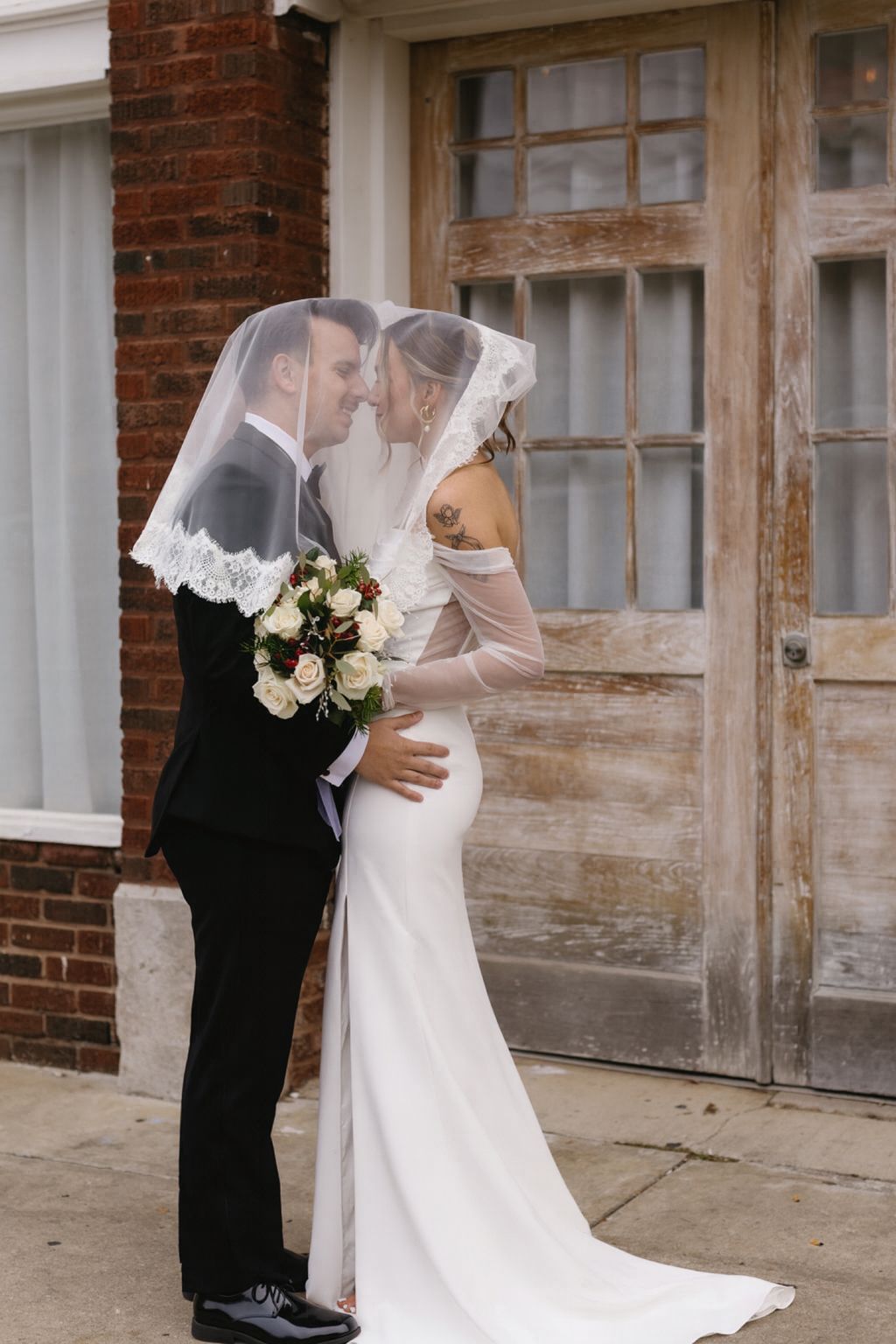 A romantic wedding couple shares a kiss against a rustic brick wall backdrop with vintage windows.
