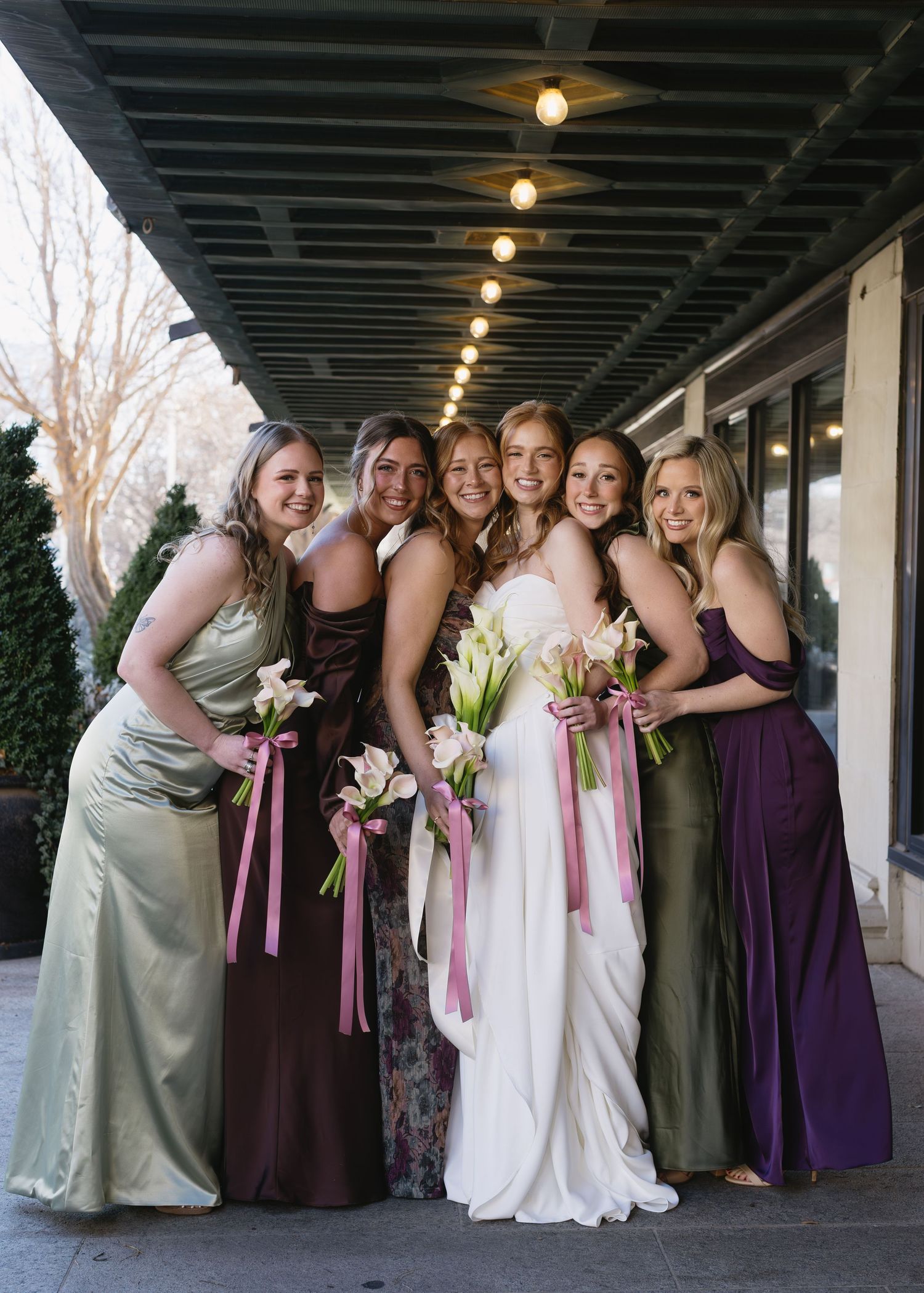 Wedding party in purple and sage dresses poses together under covered walkway.