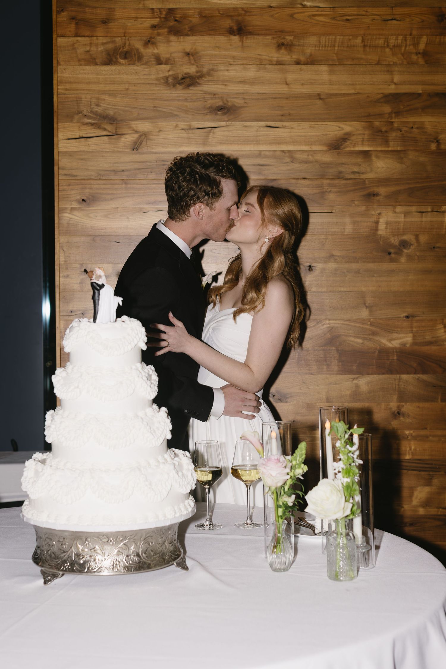 Elegant white wedding cake displayed on table with rustic wooden wall backdrop.