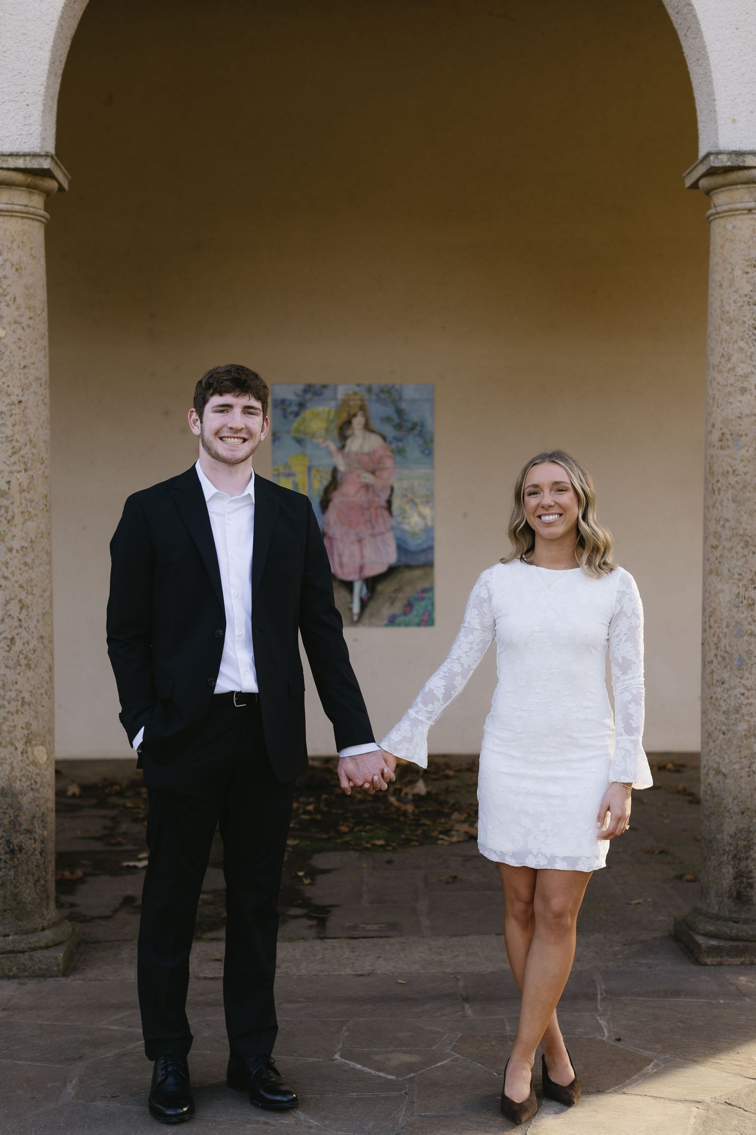 A couple poses under an archway with white columns and artwork in the background.