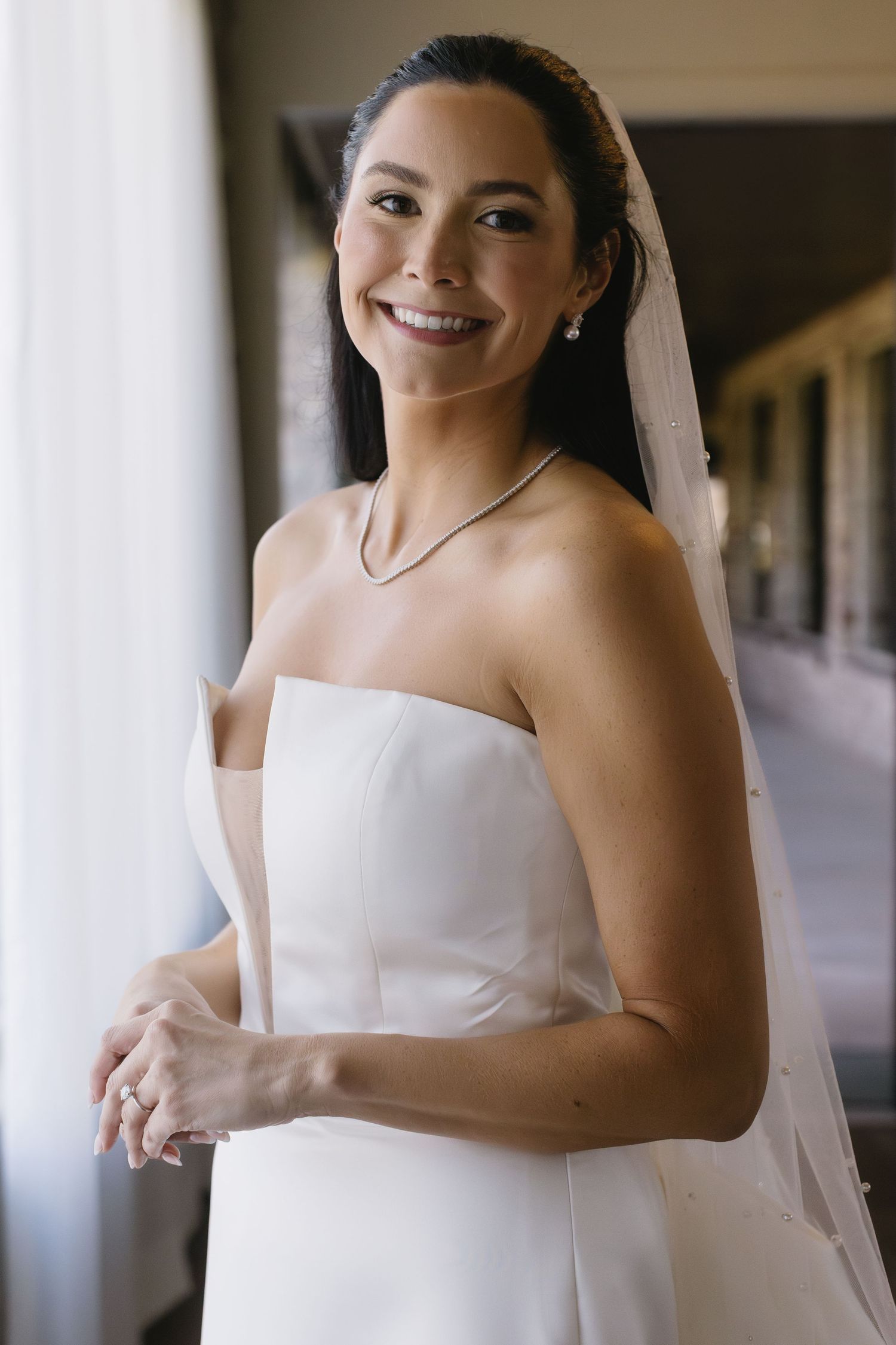 Elegant bride in strapless white wedding gown with flowing veil in indoor setting.