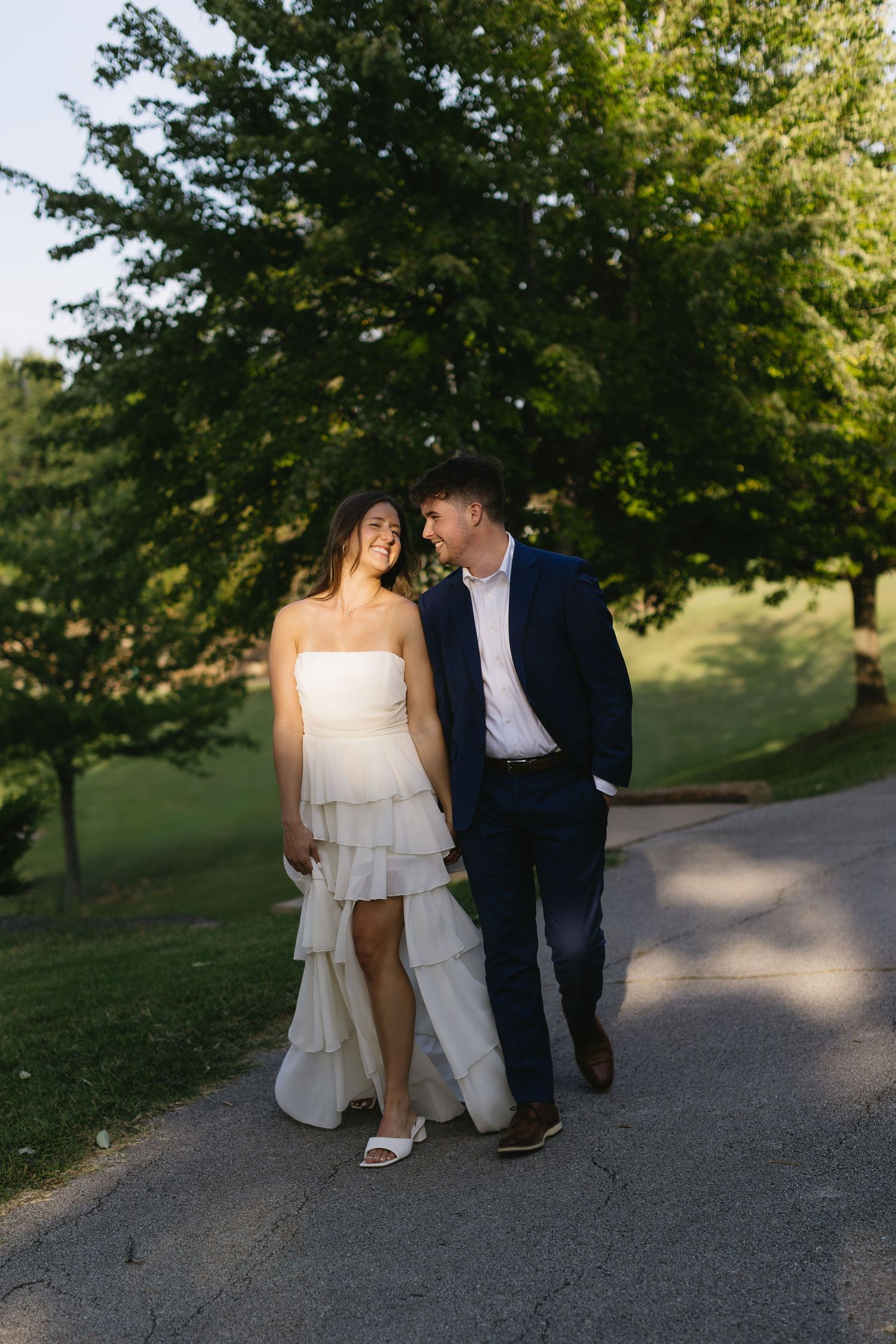 A couple walks together on a tree-lined path wearing formal attire at sunset.