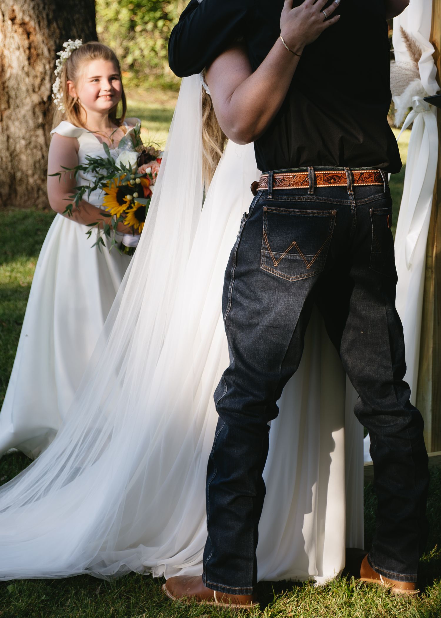 A young flower girl in white dress stands beside flowing wedding gown fabric during an outdoor ceremony.