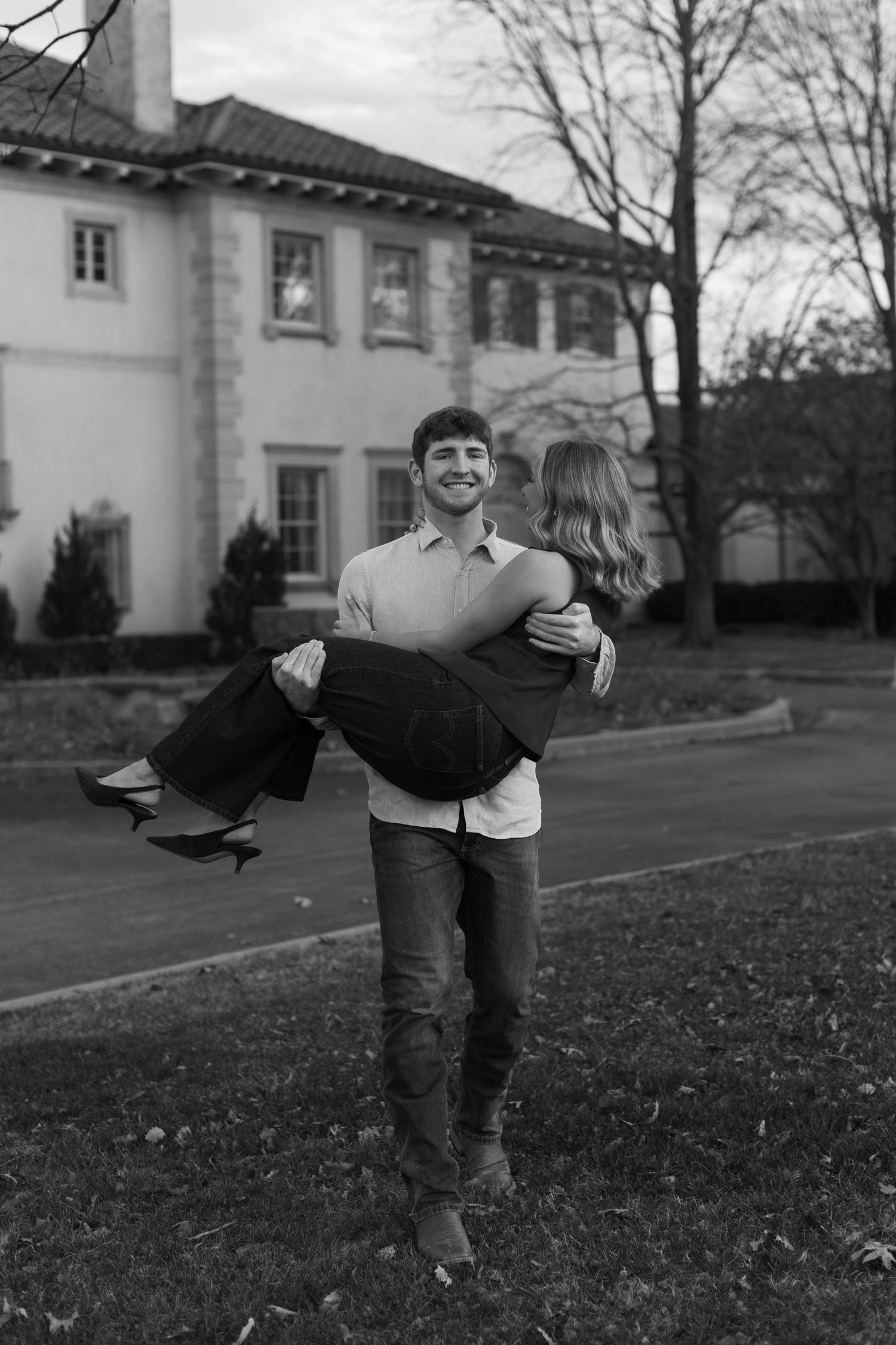 A playful black and white photo of a couple dancing on a grassy lawn near historic buildings.