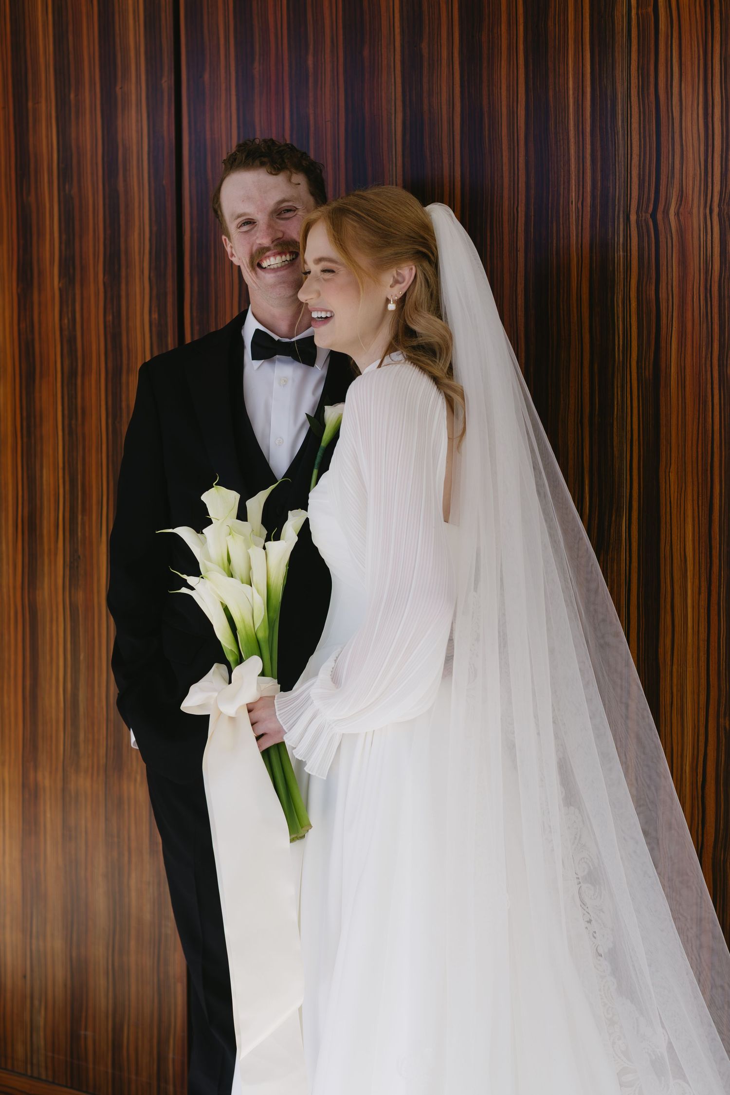 Bride in white gown holds calla lily bouquet against wood paneled wall.