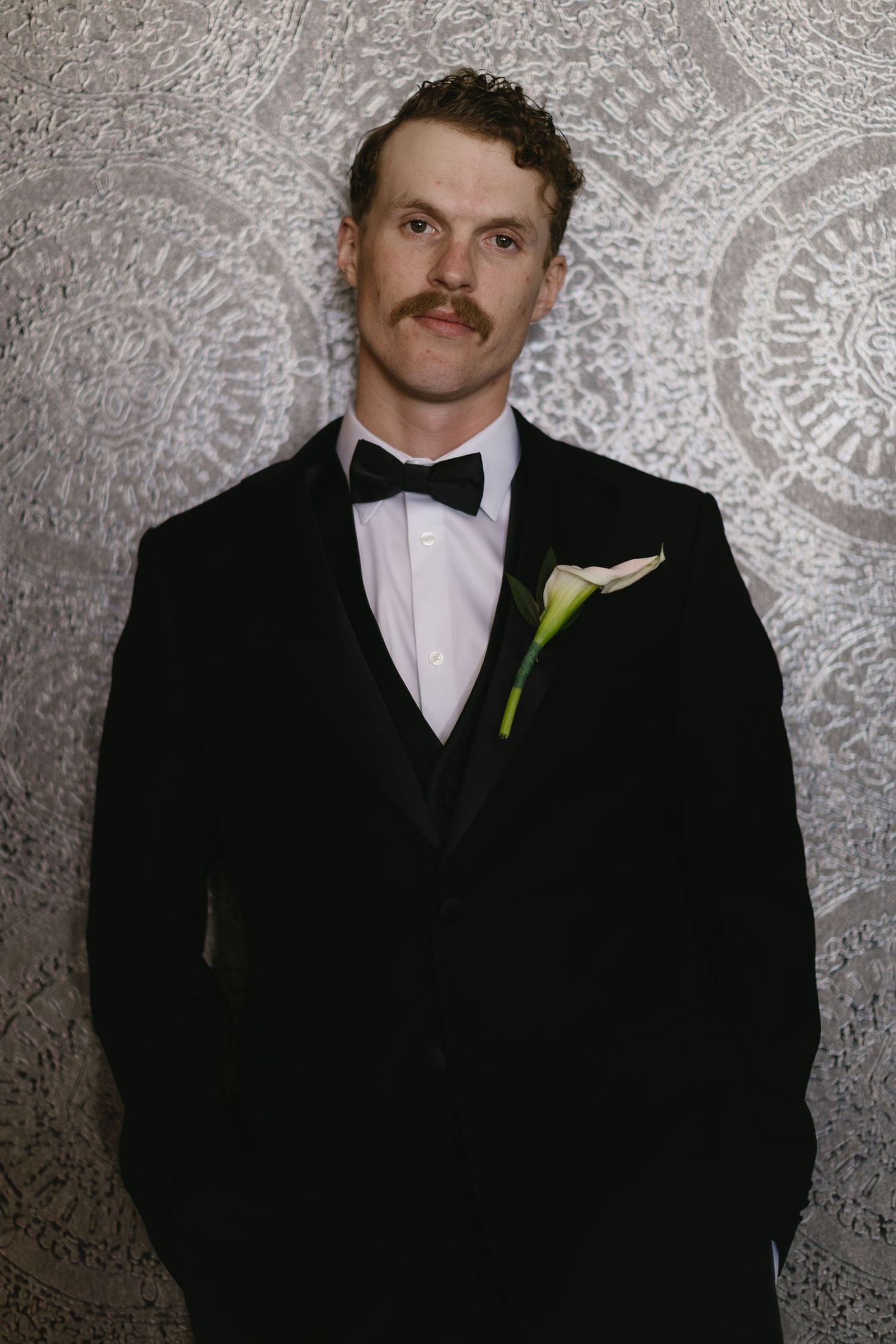A groom in a black tuxedo with a green boutonniere poses against a textured gray backdrop.