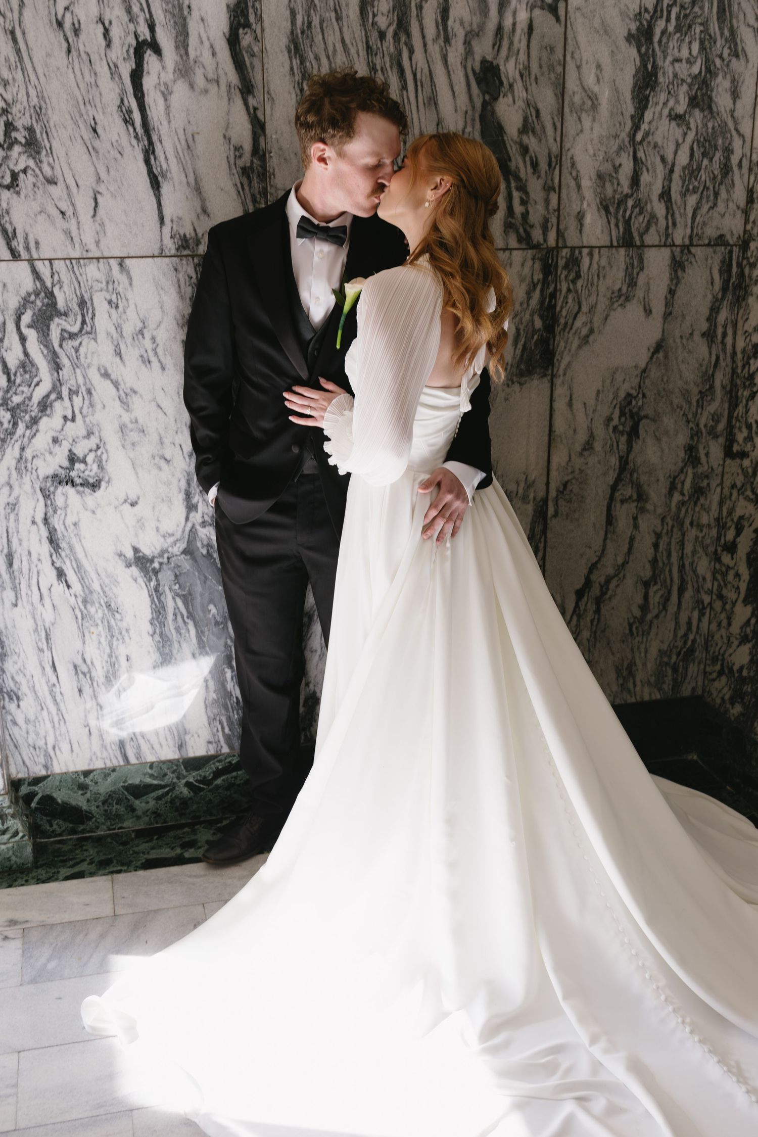 A romantic wedding couple embraces against a luxurious marble wall backdrop in an elegant venue.