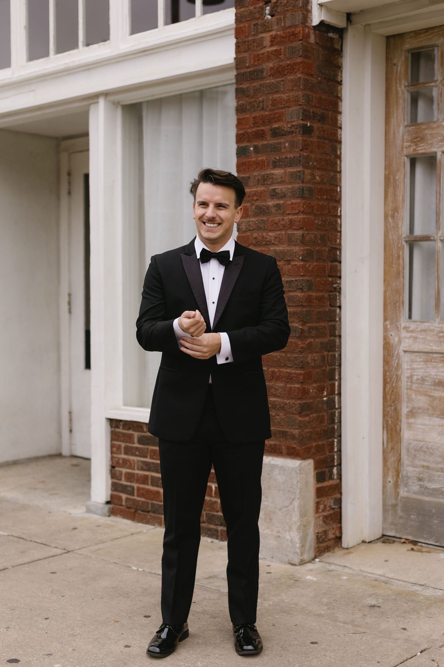 A groom in a classic black tuxedo and bow tie smiles while standing in front of a brick wall and white columns.