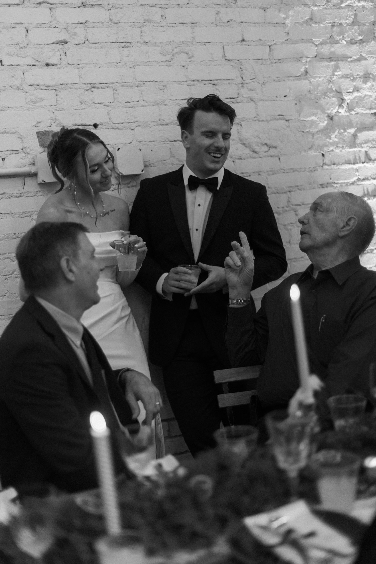 A black and white photo of wedding guests enjoying conversation at a candlelit reception table.