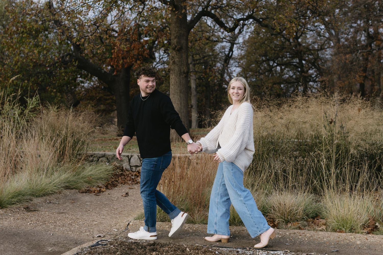 A couple walks through tall autumn grass during a romantic outdoor photo session in a natural field setting.