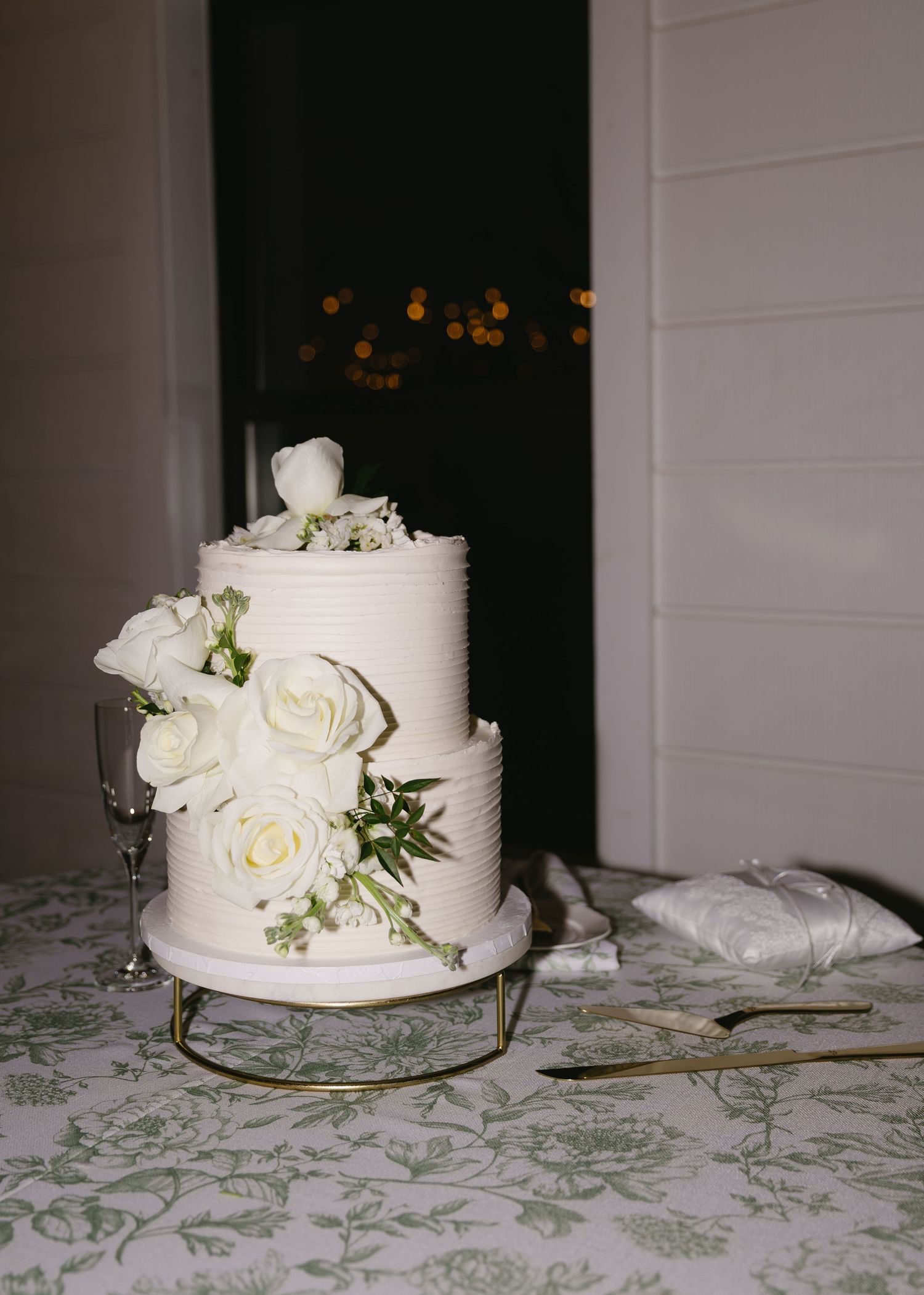 Simple white wedding cake decorated with fresh roses and greenery on a patterned tablecloth.