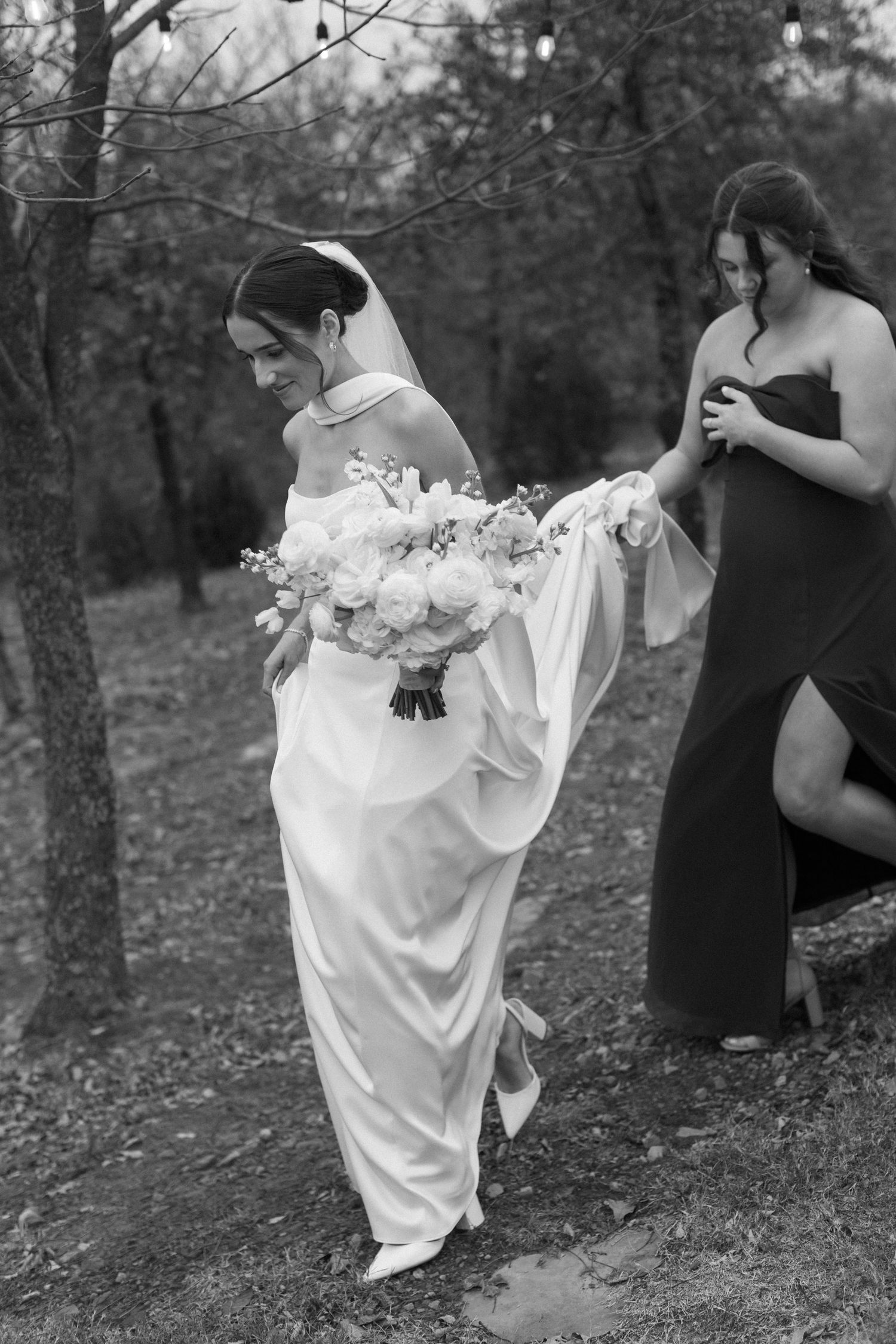 A bride in a white dress lifts her gown while walking through autumn leaves with bridesmaids in black dresses.