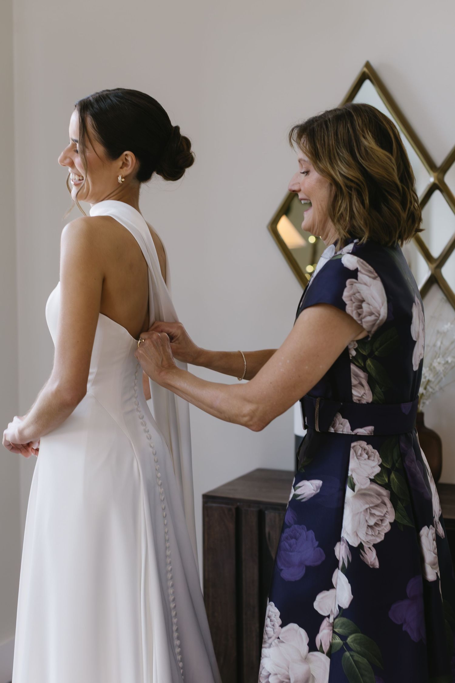 A bride in a white gown gets help with dress buttons from someone wearing a navy floral dress.
