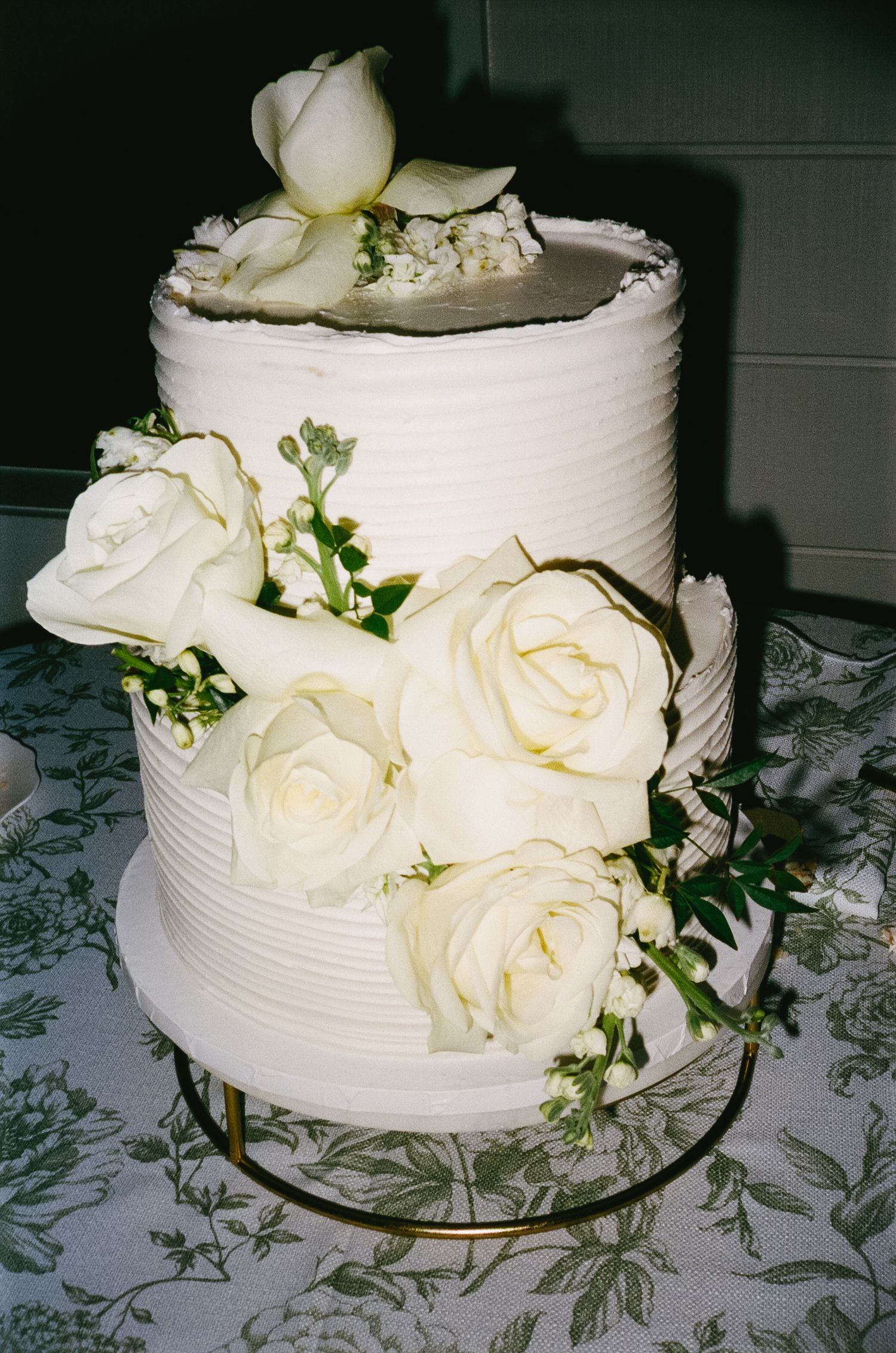 White wedding cake decorated with fresh cream roses and greenery on a patterned tablecloth.