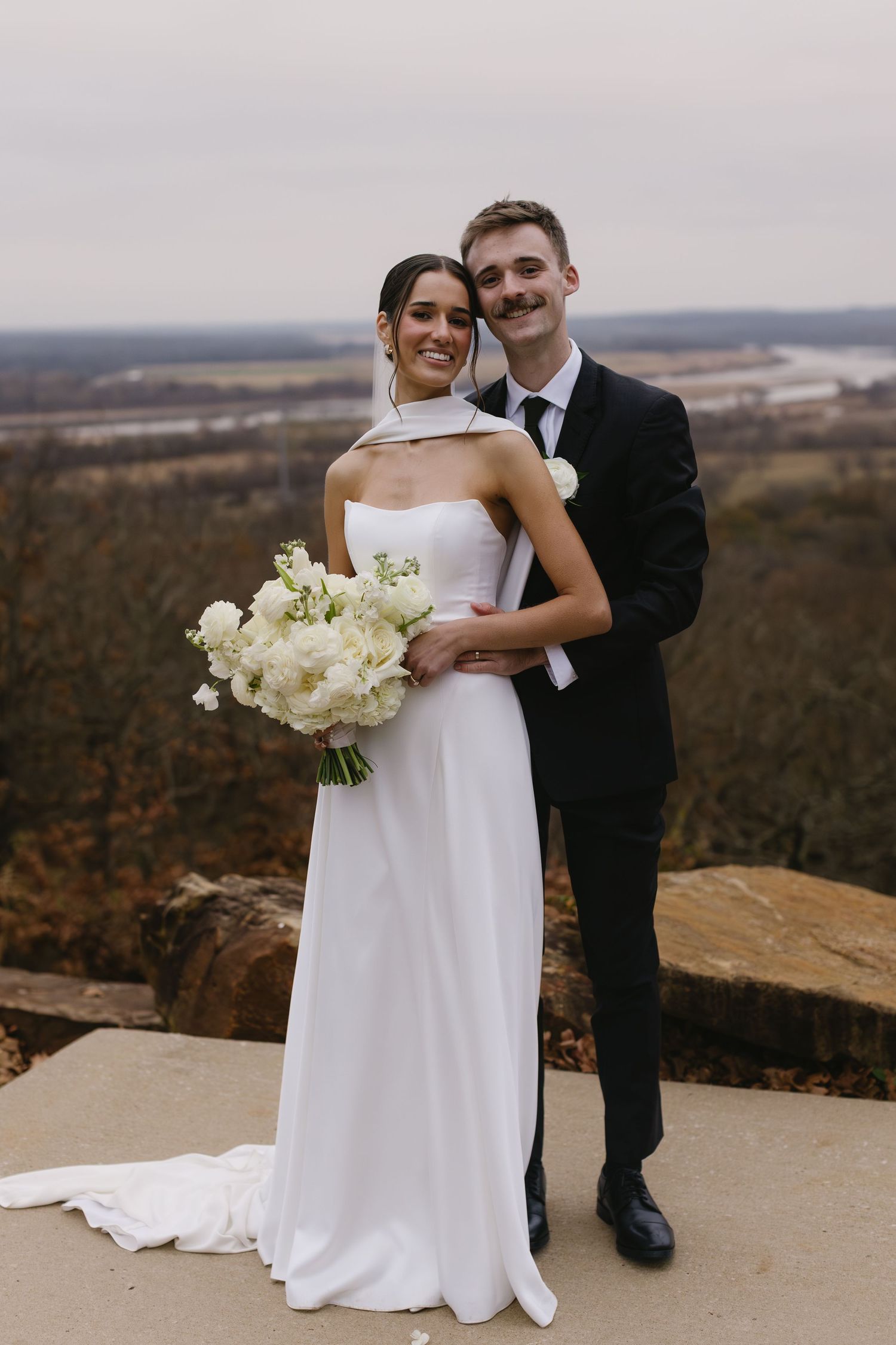 Wedding couple shares intimate moment on scenic overlook with bride holding white bouquet.