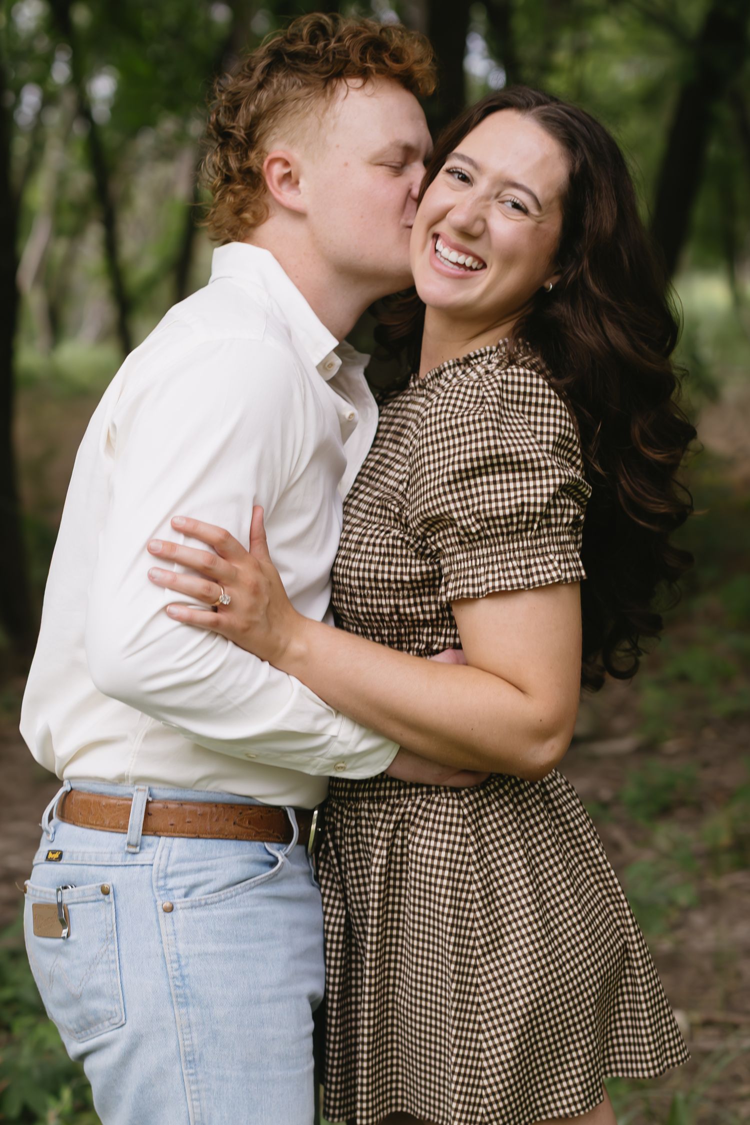 A couple in vintage-inspired outfits embrace playfully in a wooded setting.
