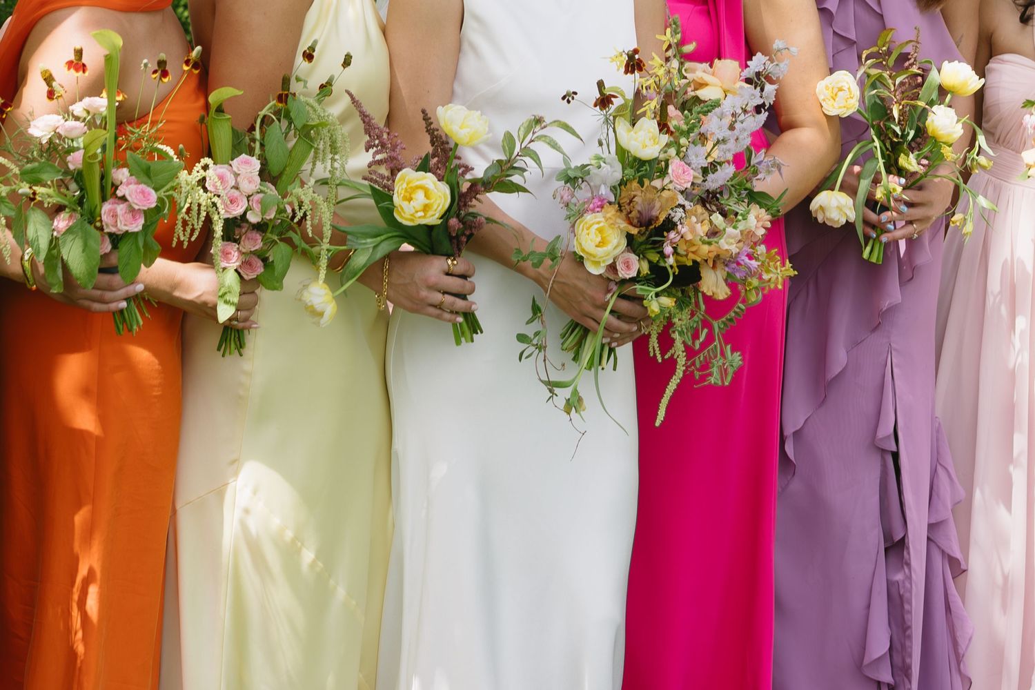 Bridesmaids holding floral bouquets while wearing colorful dresses in orange, pink, and purple shades.