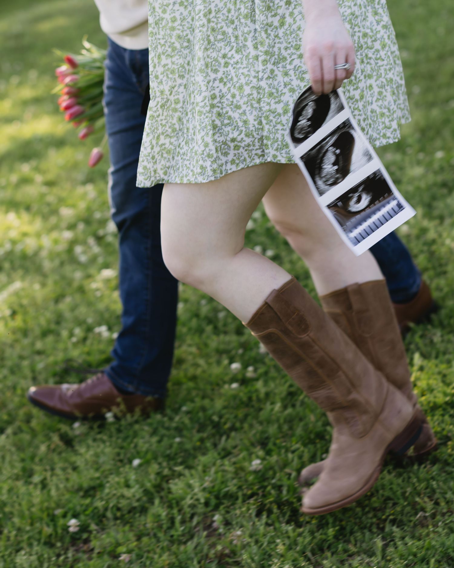 Close-up of brown boots and floral dress against green grass with ultrasound photos visible.