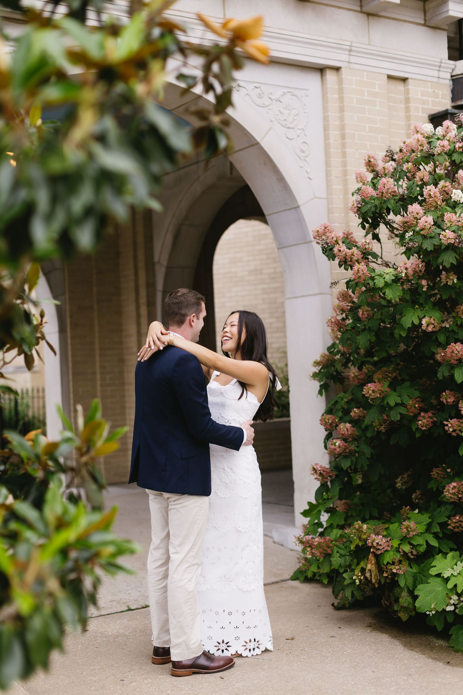 Wedding couple embraces and dances in front of a white columned building with flowering bushes.