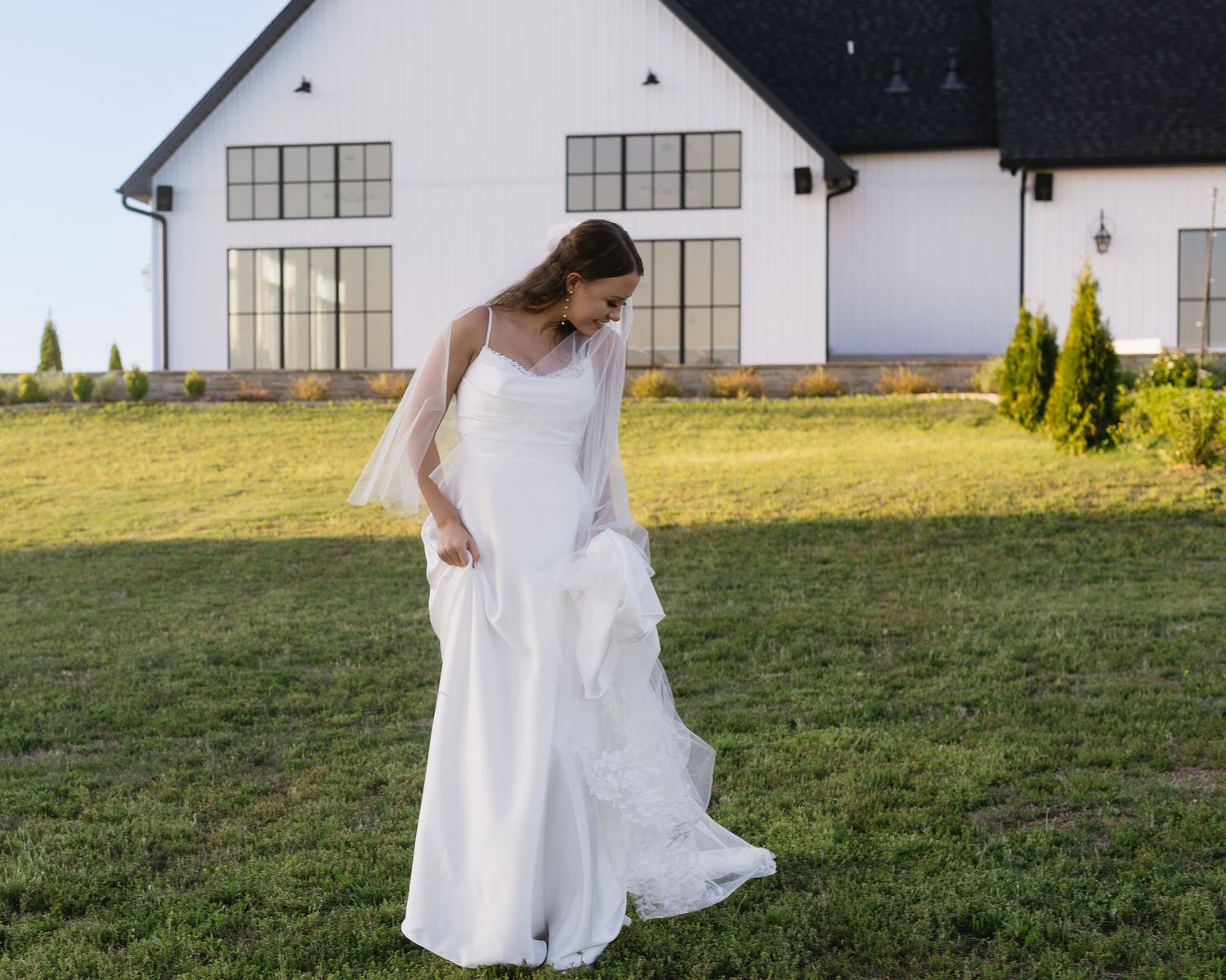 Wedding dress swirls against backdrop of white barn buildings and green grass.